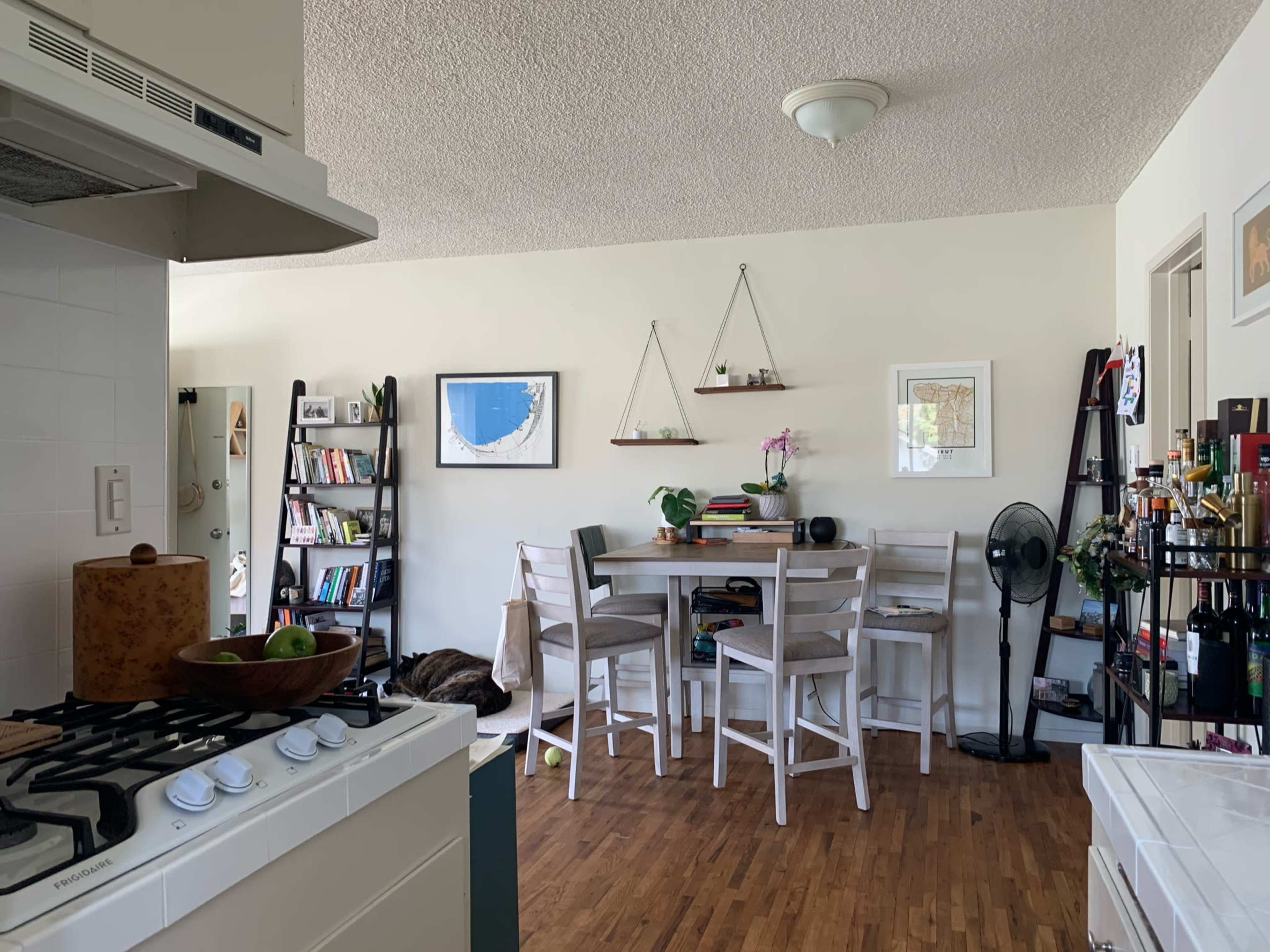 The image shows a bright, open-concept living space with a kitchen on the left, featuring a gas stove, and a dining area with a table and chairs, along with bookshelves and decorative plants in the background.