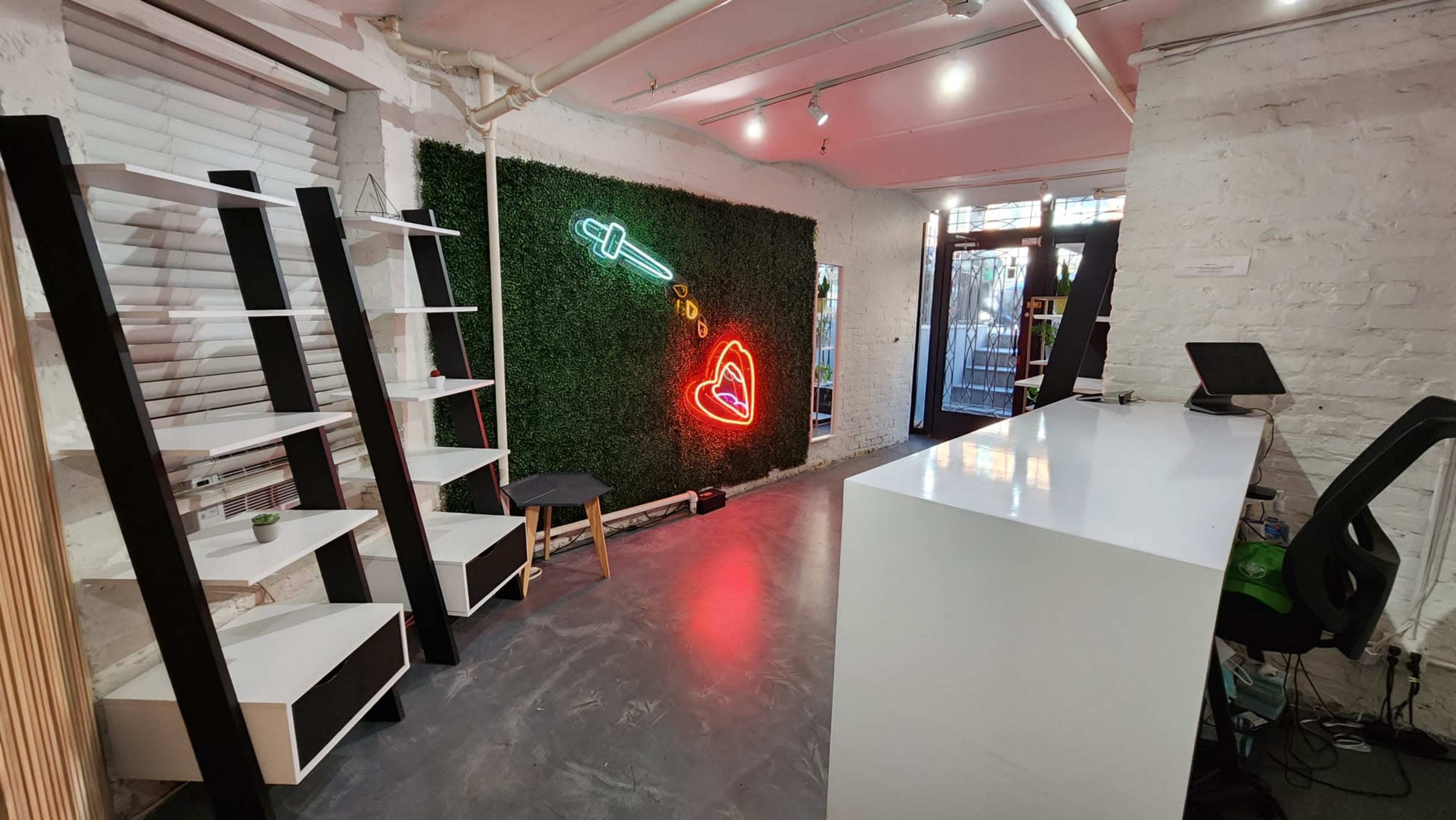 The scene features a minimalist interior with white walls, a reception desk, shelves, and a neon heart and syringe design on a green wall.