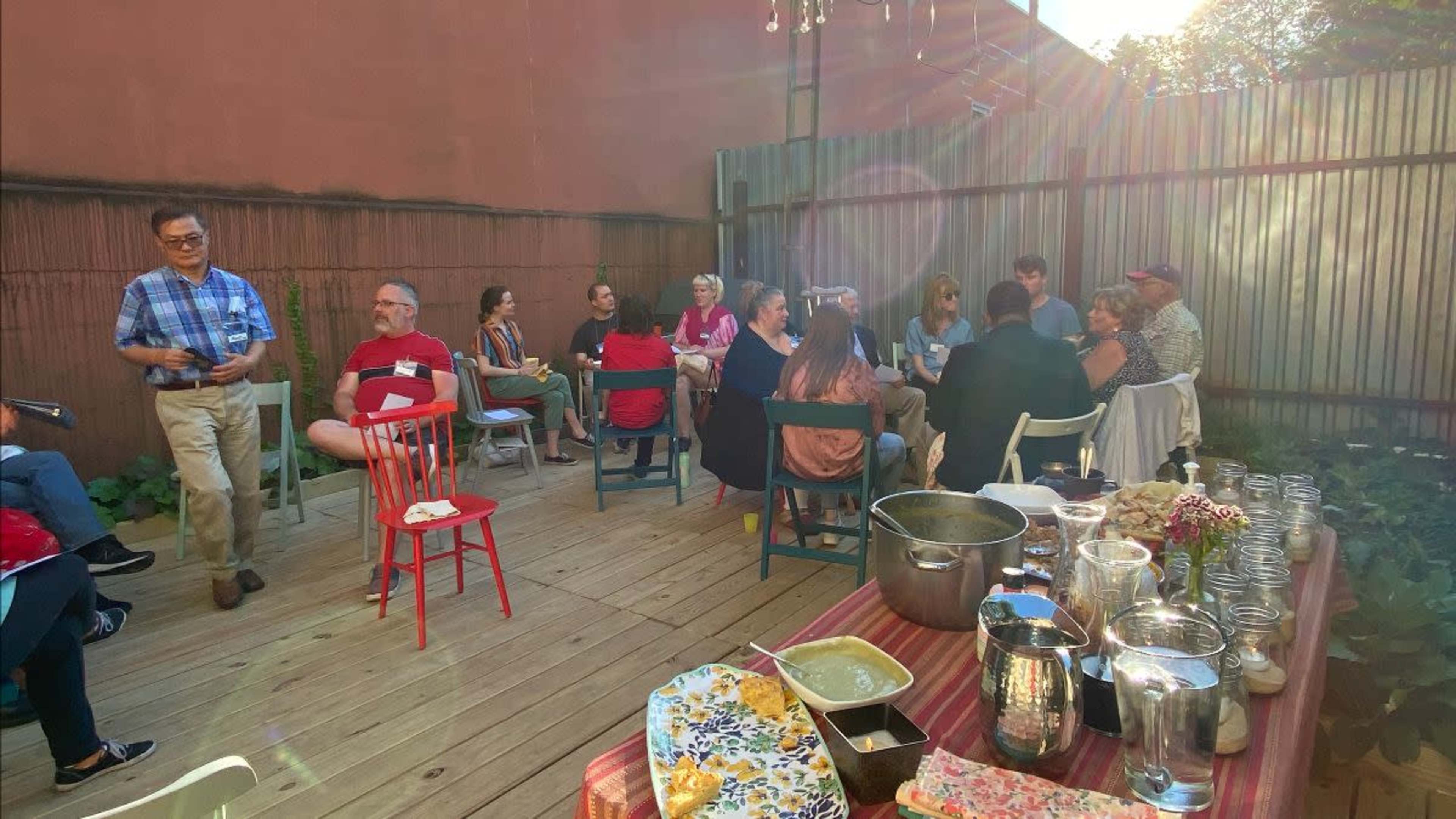 A group of people is seated in a courtyard, engaged in conversation, with several tables displaying food and drinks.