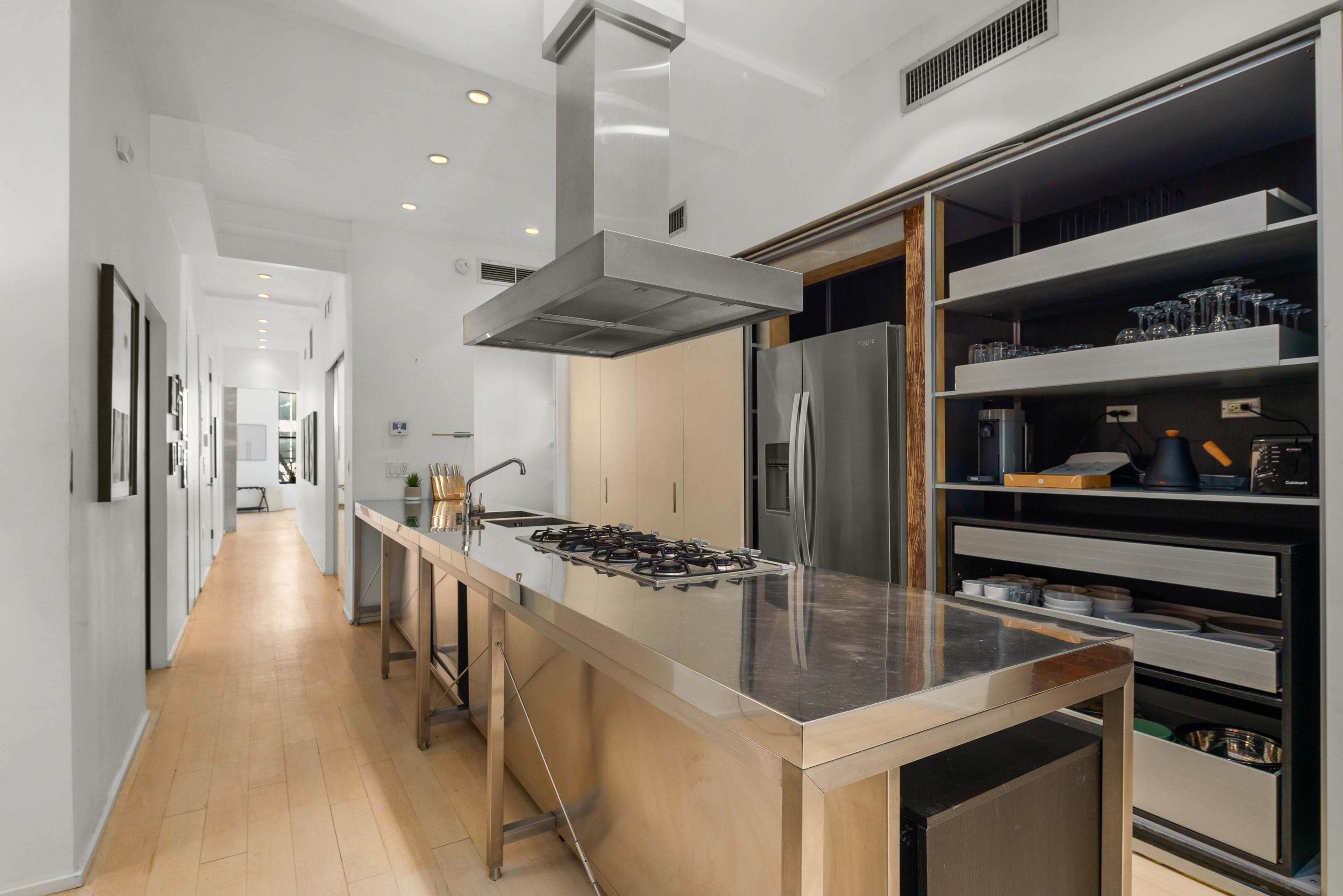 A modern kitchen features a central stainless steel island with a gas stove and overhead ventilation, complemented by sleek cabinetry and a long hallway leading to another room.