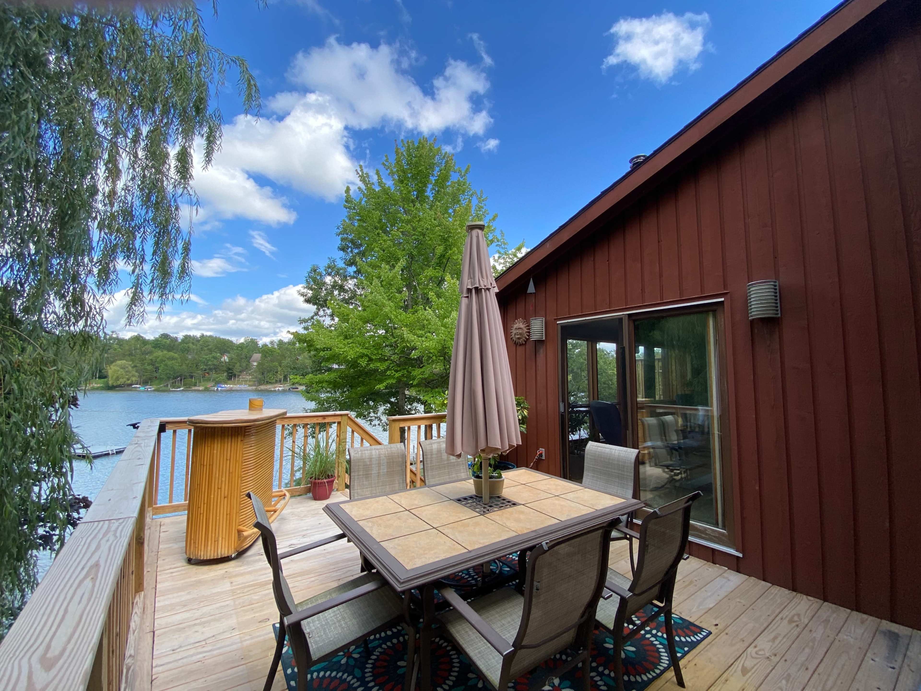 A wooden deck with a table and chairs is set up by a lake, surrounded by greenery and under a partly cloudy sky.