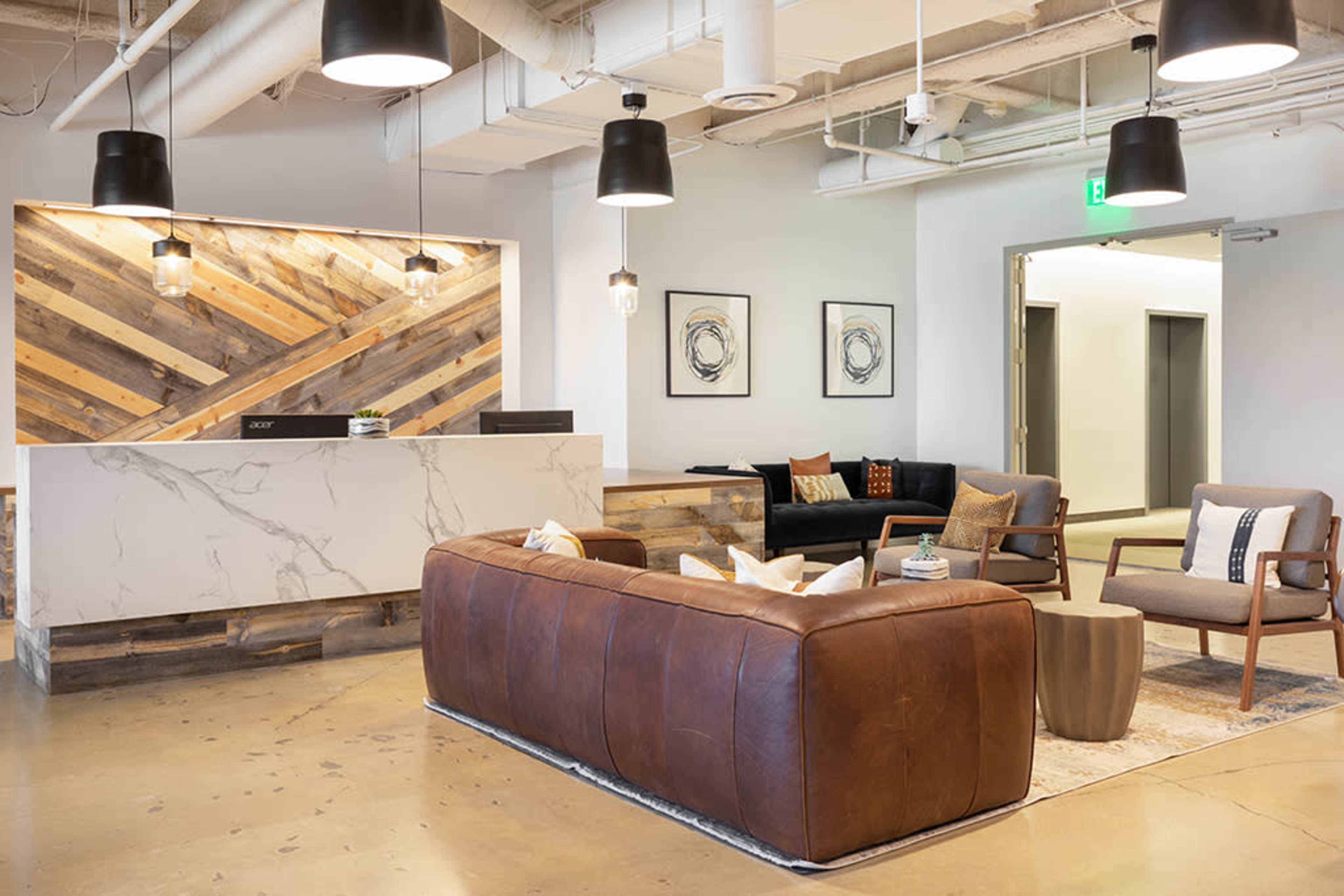 A modern reception area with a seating arrangement, featuring a dark leather sofa, two armchairs, and a front desk with a wooden backdrop.