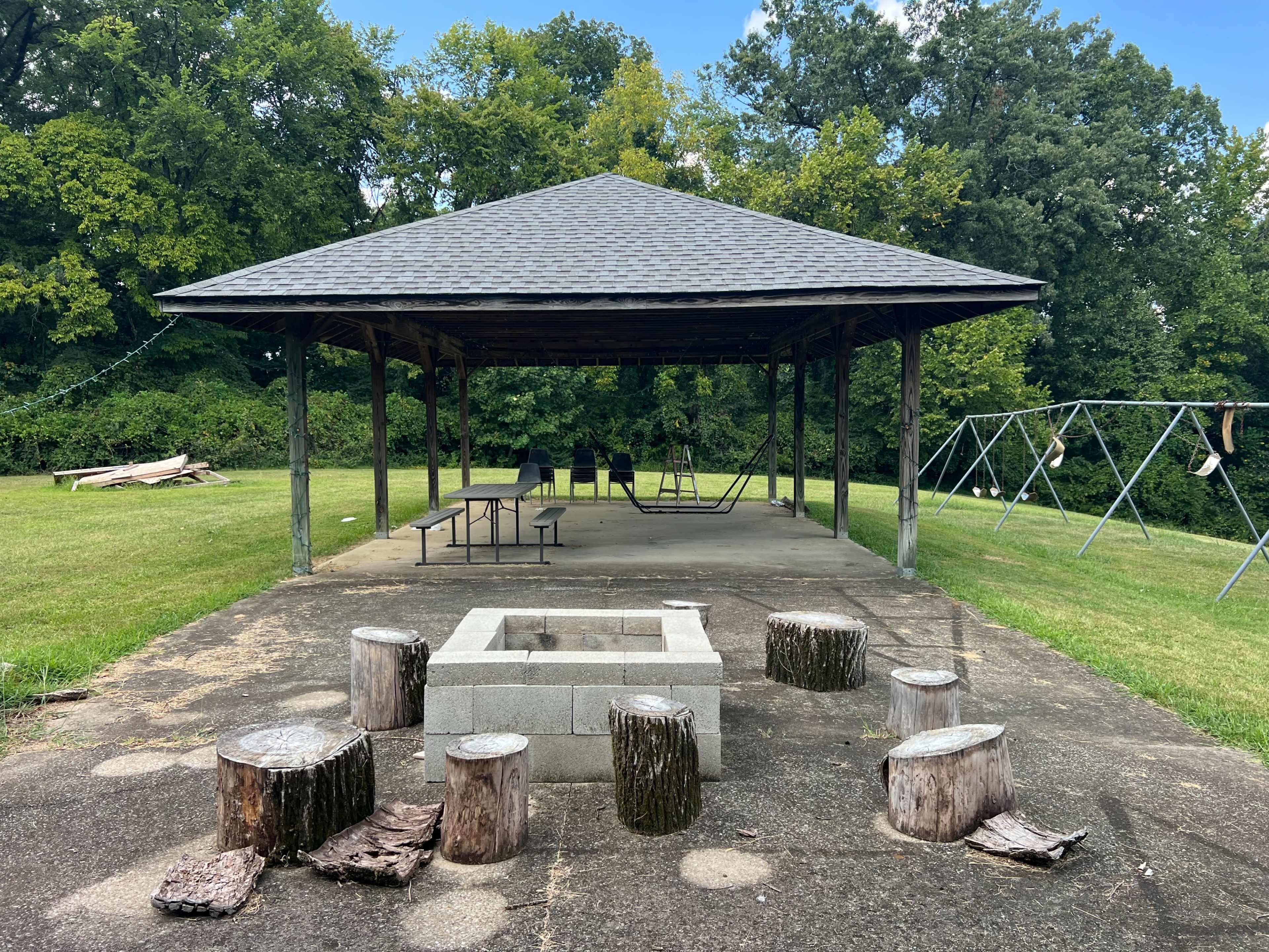 The image shows a shaded pavilion with picnic tables and a fire pit surrounded by logs, set in a grassy area near trees.