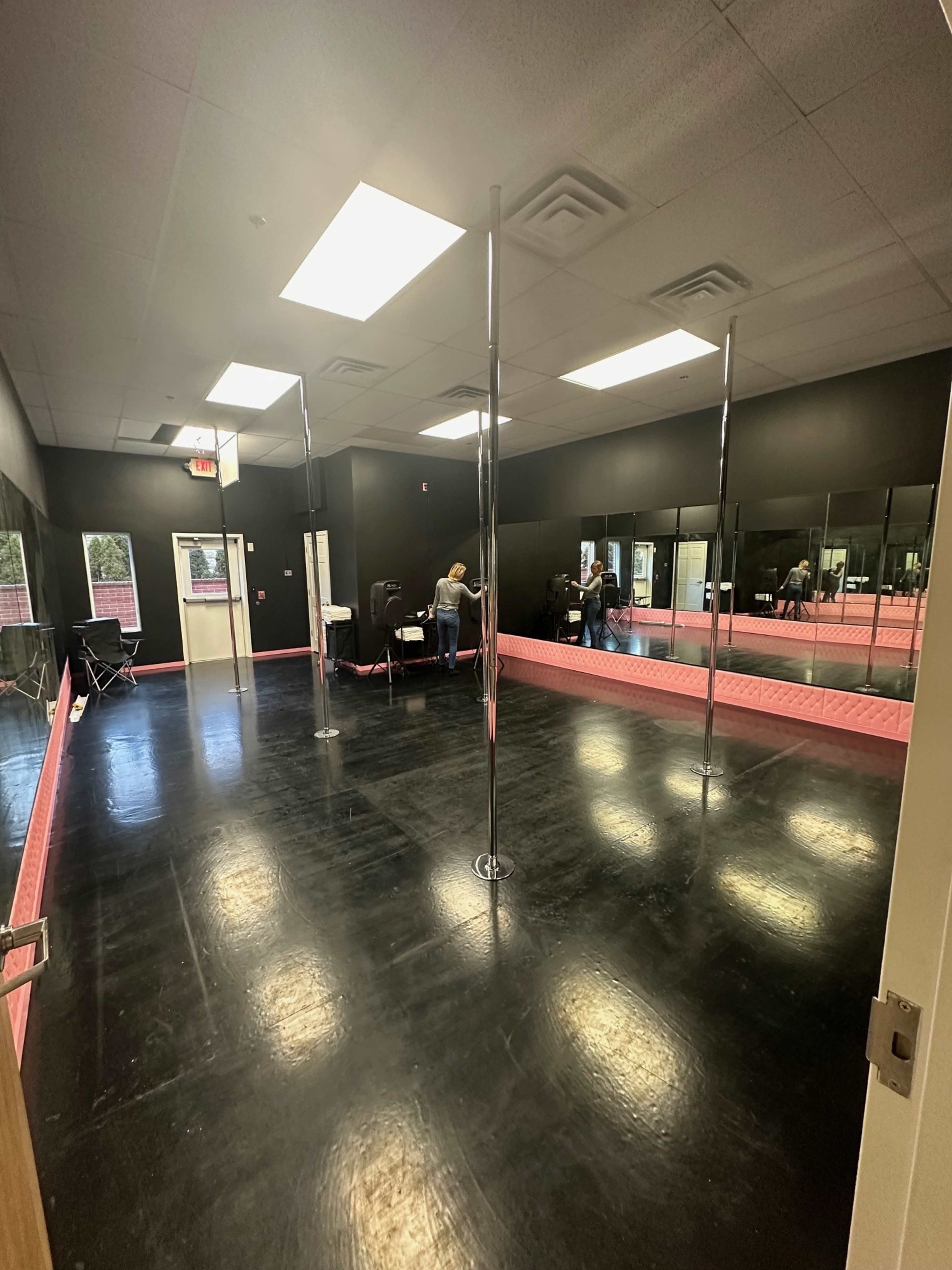 A spacious dance studio with polished black floors, several pole dance apparatuses, and mirrored walls.