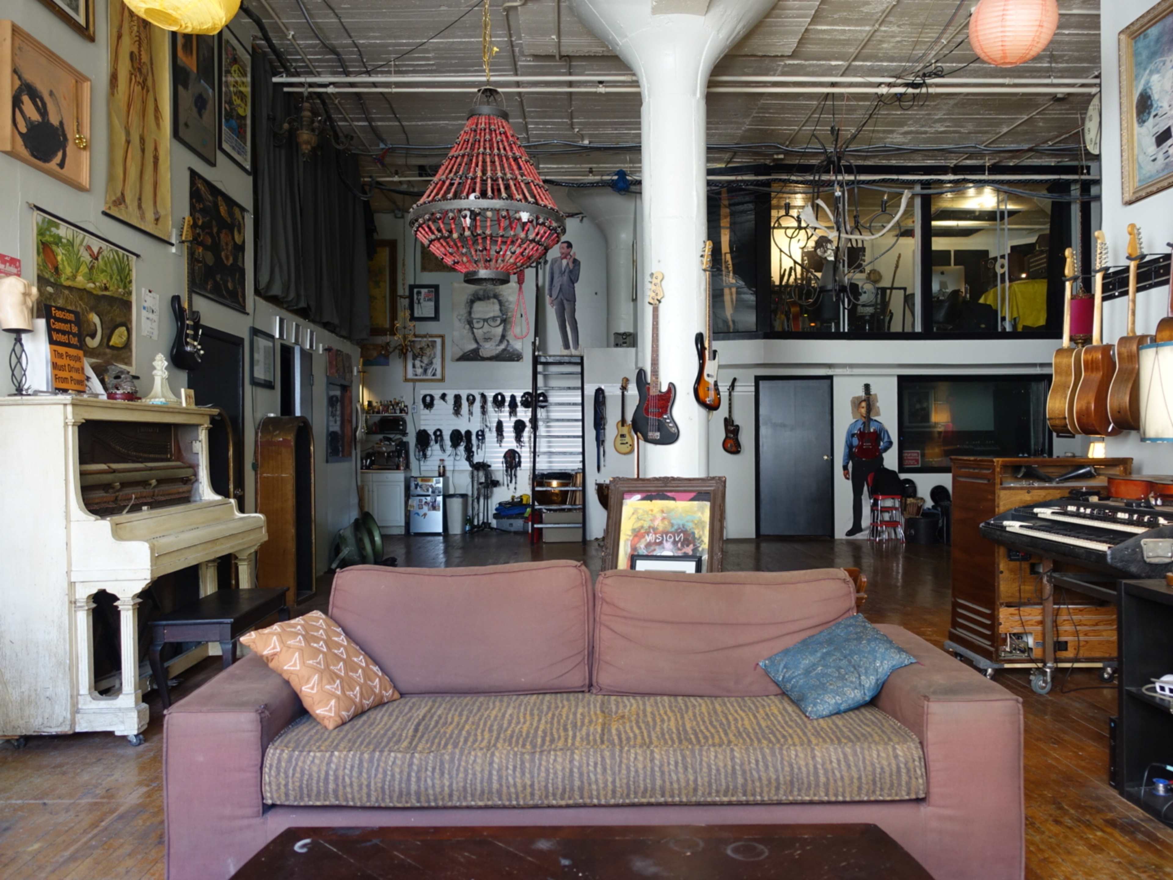 The image shows a spacious, eclectic room featuring a brown sofa, musical instruments, artwork on the walls, and a piano.