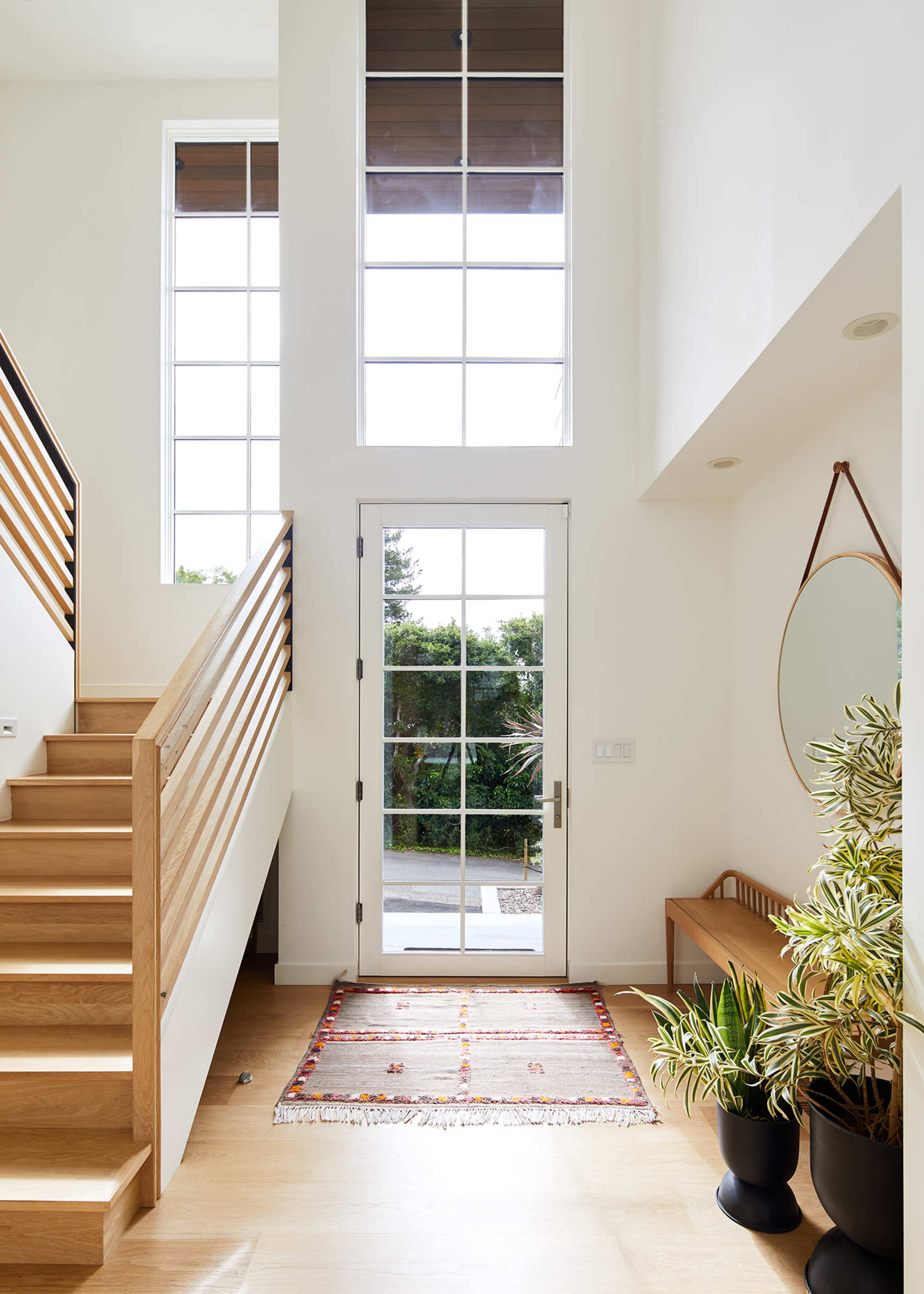 The image shows a bright entryway with a staircase on the left, a door with large windows, and plants in the corner.