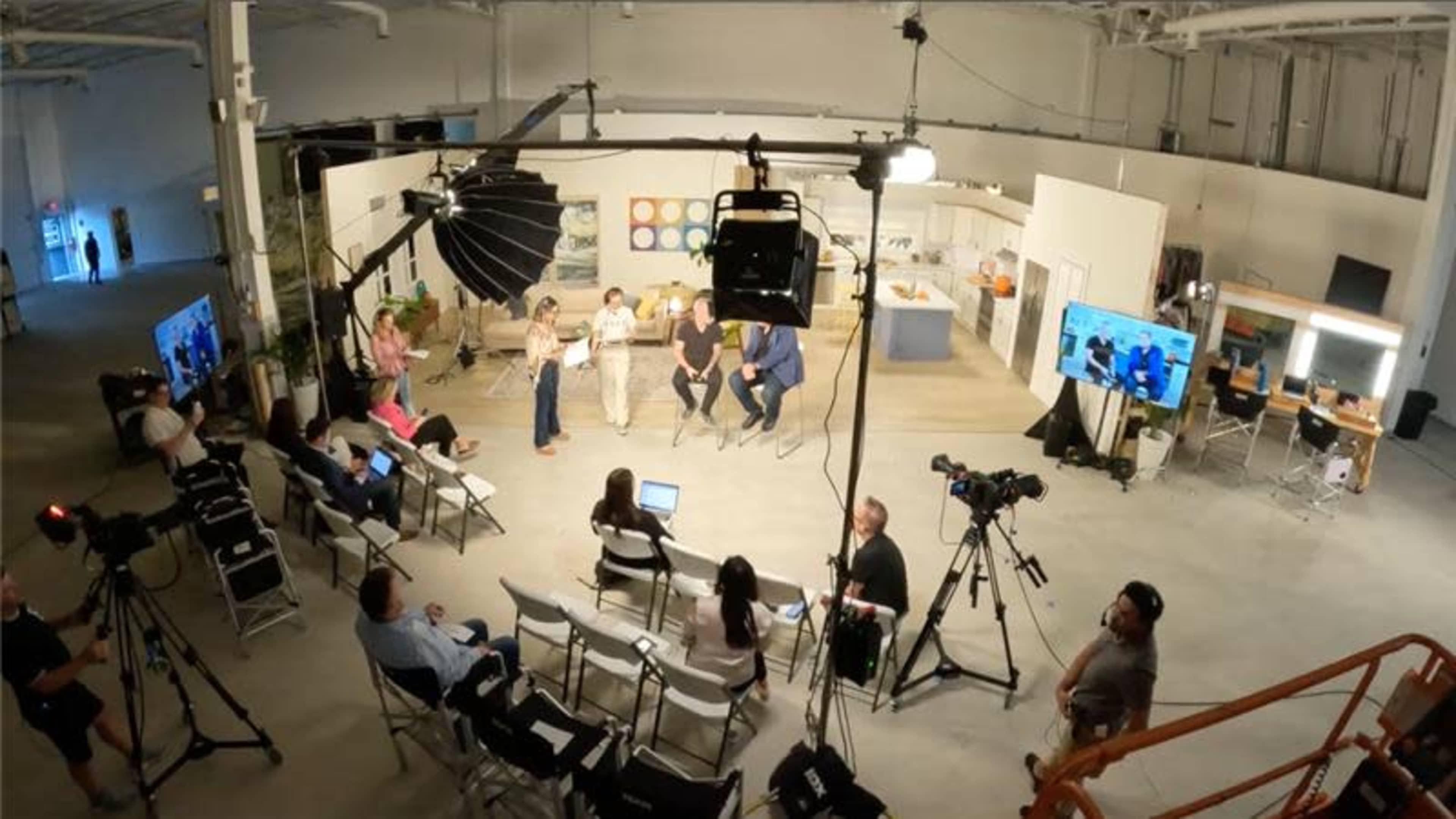 A group of people sits on a stage in a well-lit studio while cameras and audience members observe from folding chairs.