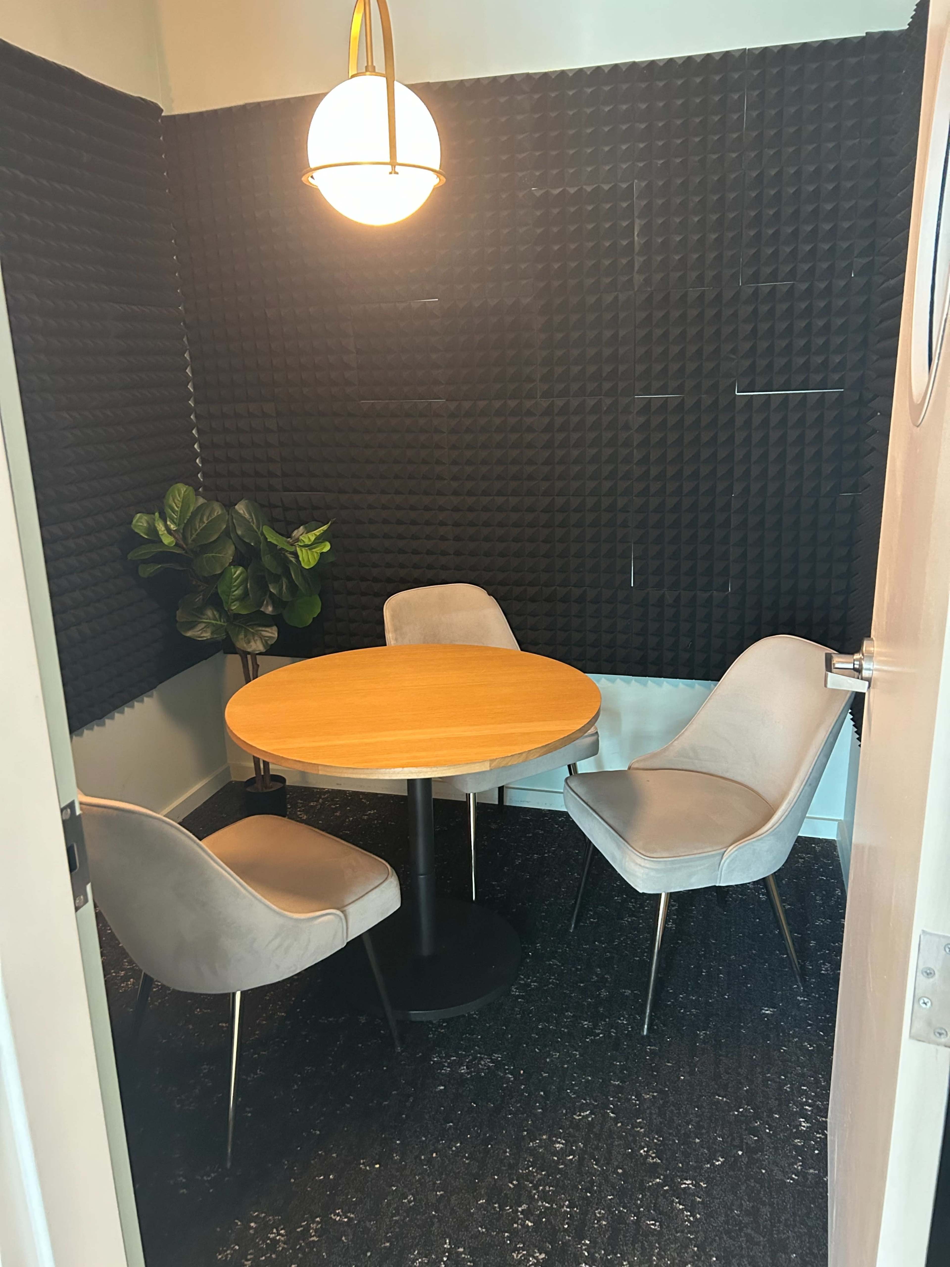 A small meeting room features a circular wooden table surrounded by three gray chairs, with black acoustic panels on the walls and a hanging light fixture above.