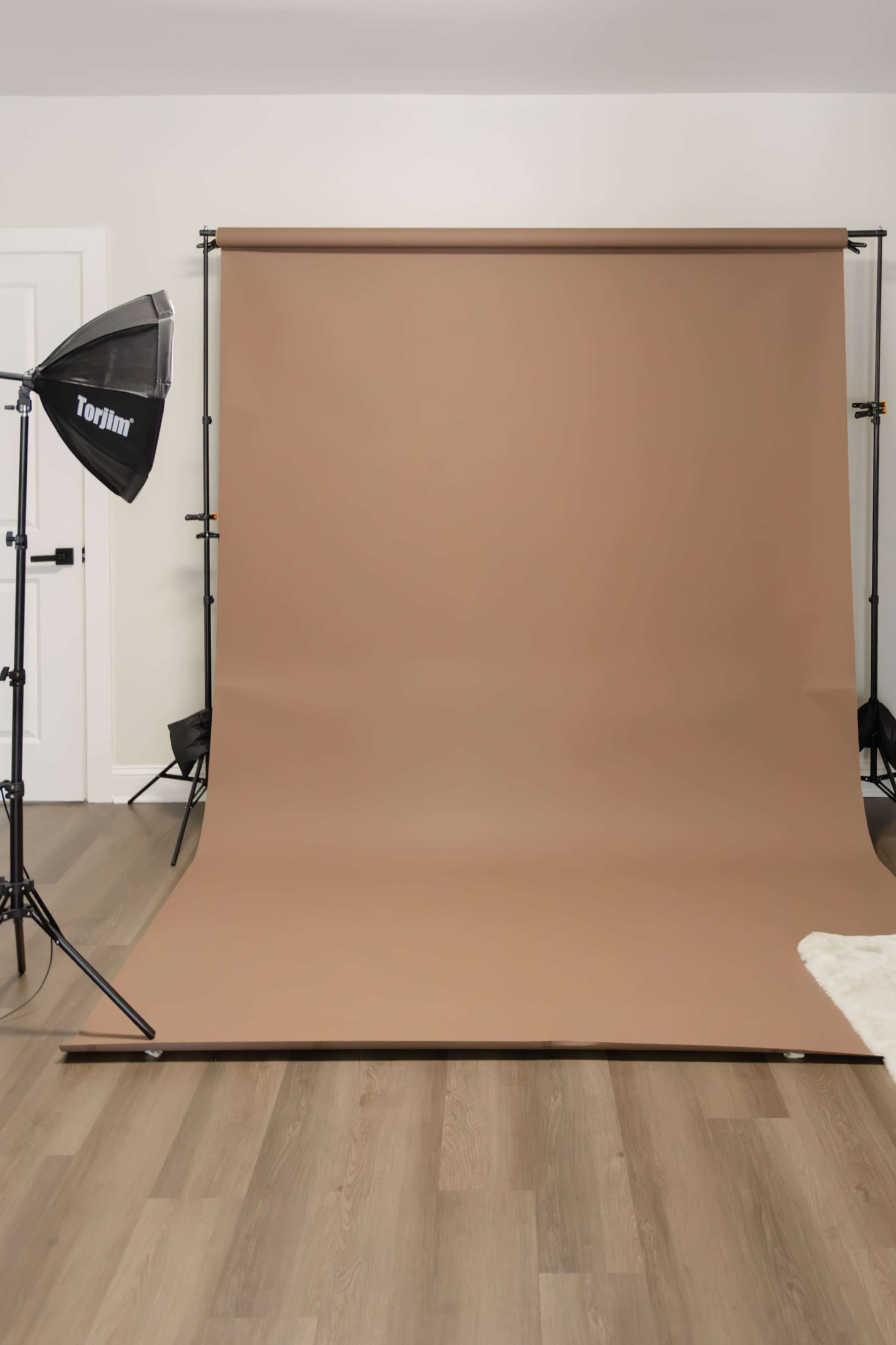 The image shows a photography studio setup with a brown backdrop, a softbox light on a stand, and wooden flooring.