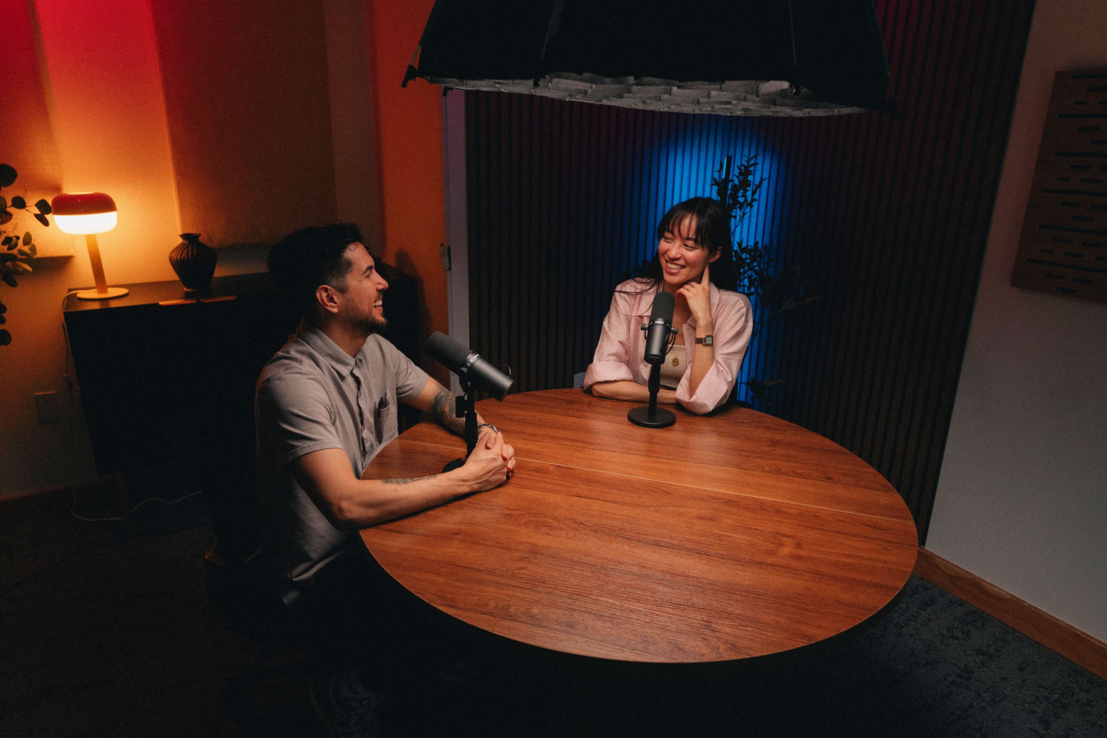 Two individuals sit across from each other at a round wooden table with microphones in a well-lit room featuring colorful ambient lighting.
