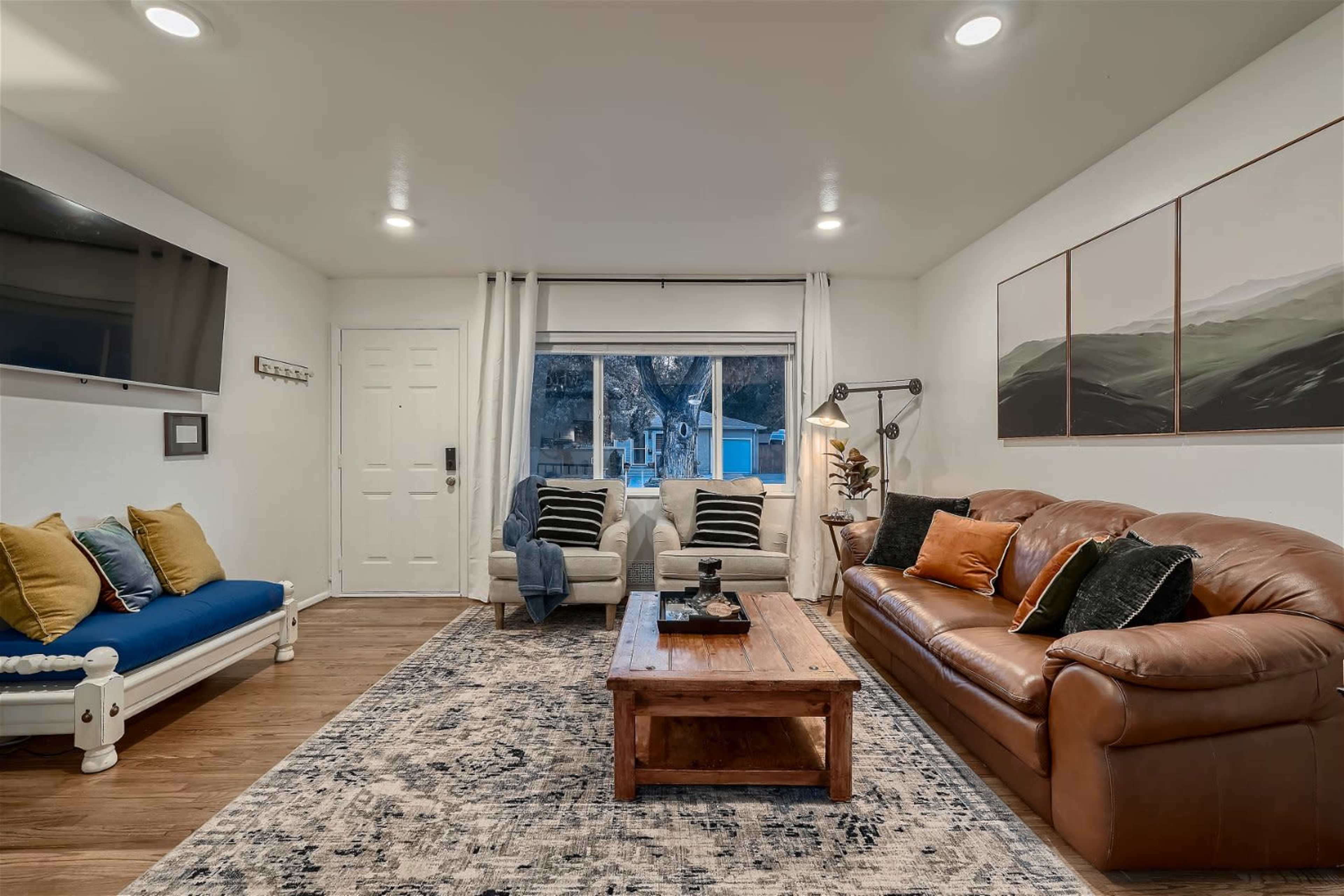 The image shows a living room featuring a brown leather sofa, a wooden coffee table, two armchairs, and a large window with natural light, adorned with decorative pillows and artwork on the walls.