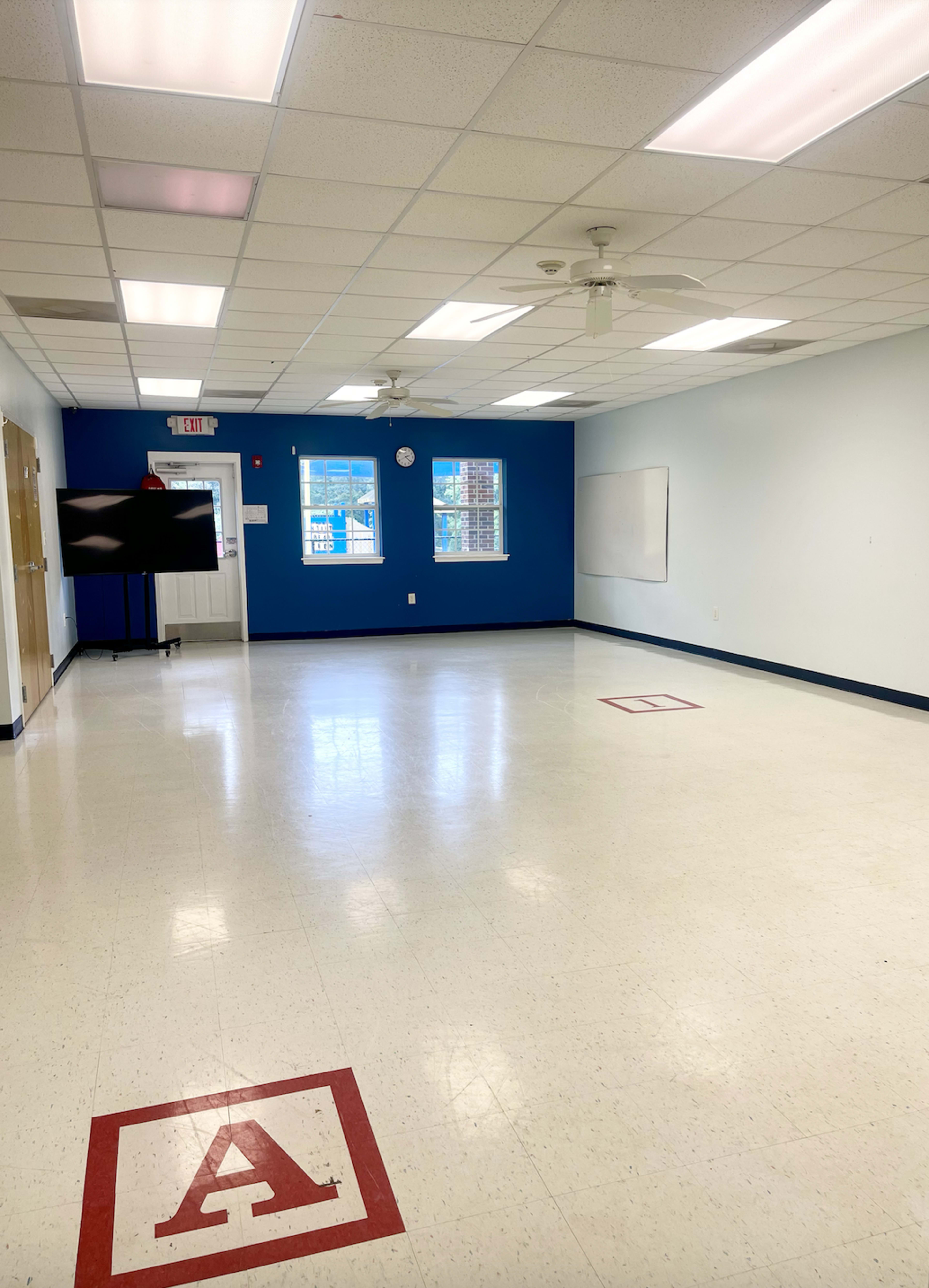 The image shows a spacious, empty classroom with blue and light blue walls, a whiteboard, and a large screen against one wall.