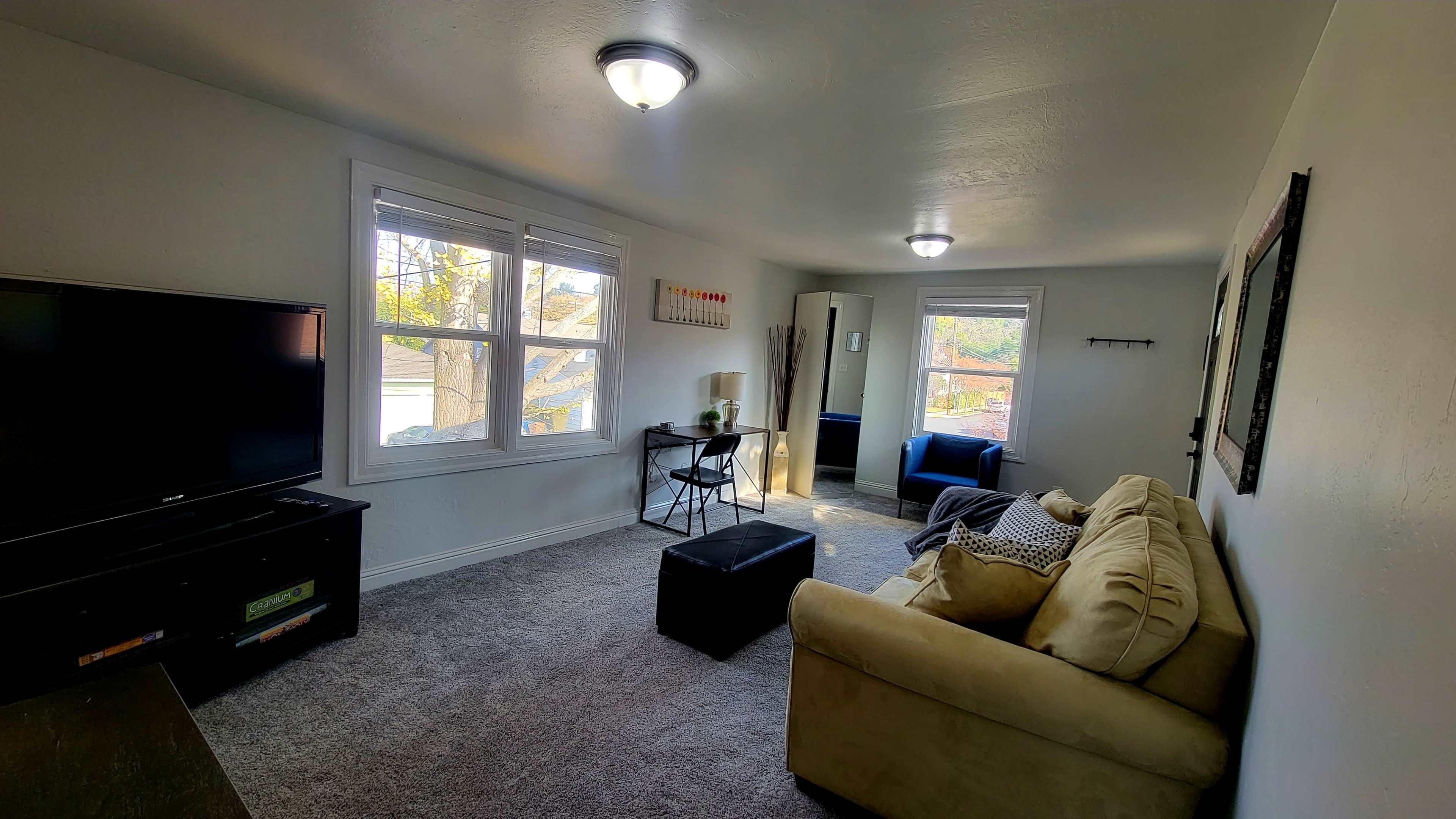The living room features a beige couch, a black ottoman, a television stand, and two blue chairs, with natural light coming through the windows.