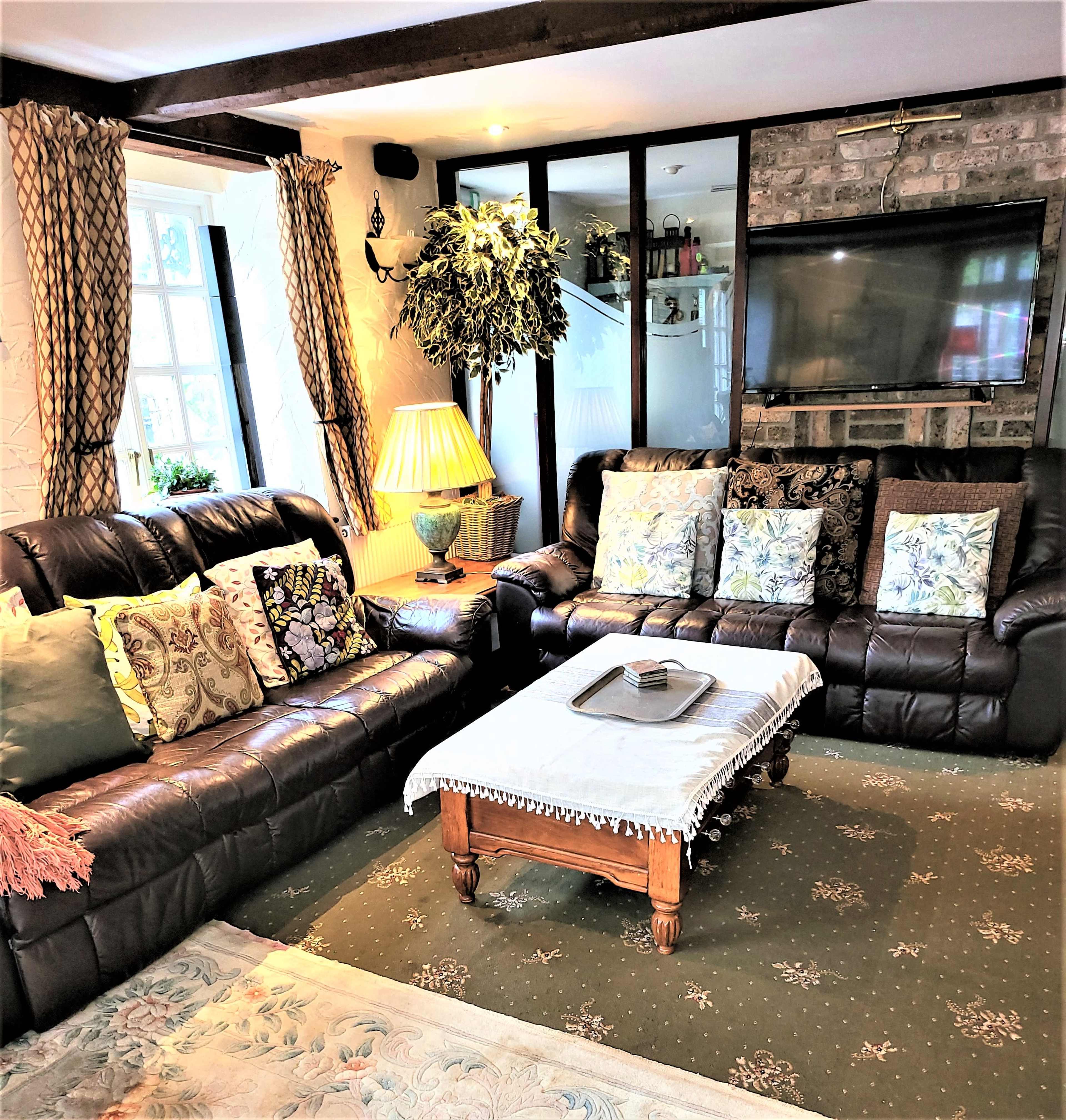 The image shows a cozy living room featuring two dark leather sofas adorned with decorative cushions, a wooden coffee table at the center, and a potted plant beside a lamp.