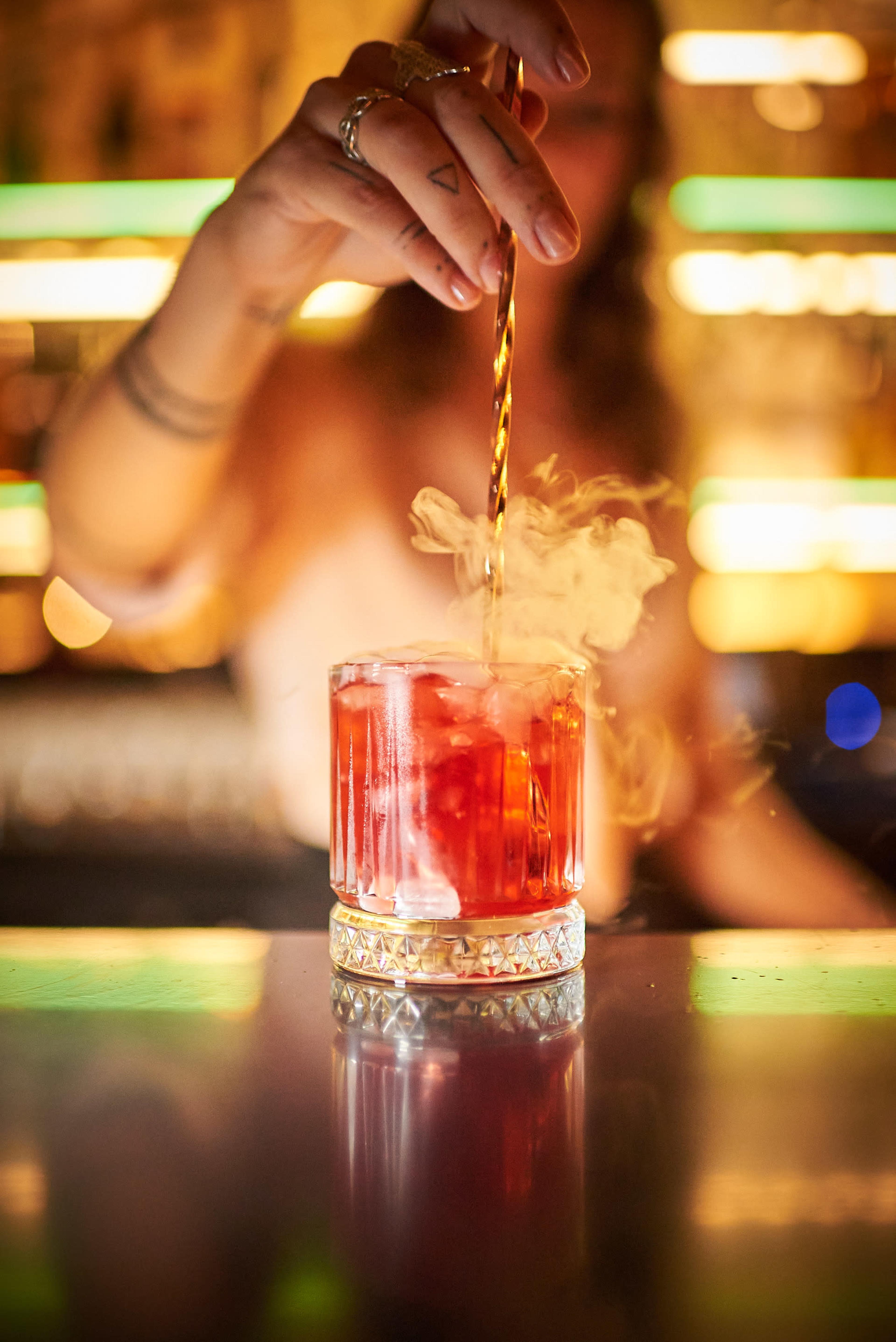 A bartender stirs a cocktail in a glass with ice, while vapor rises from the drink.