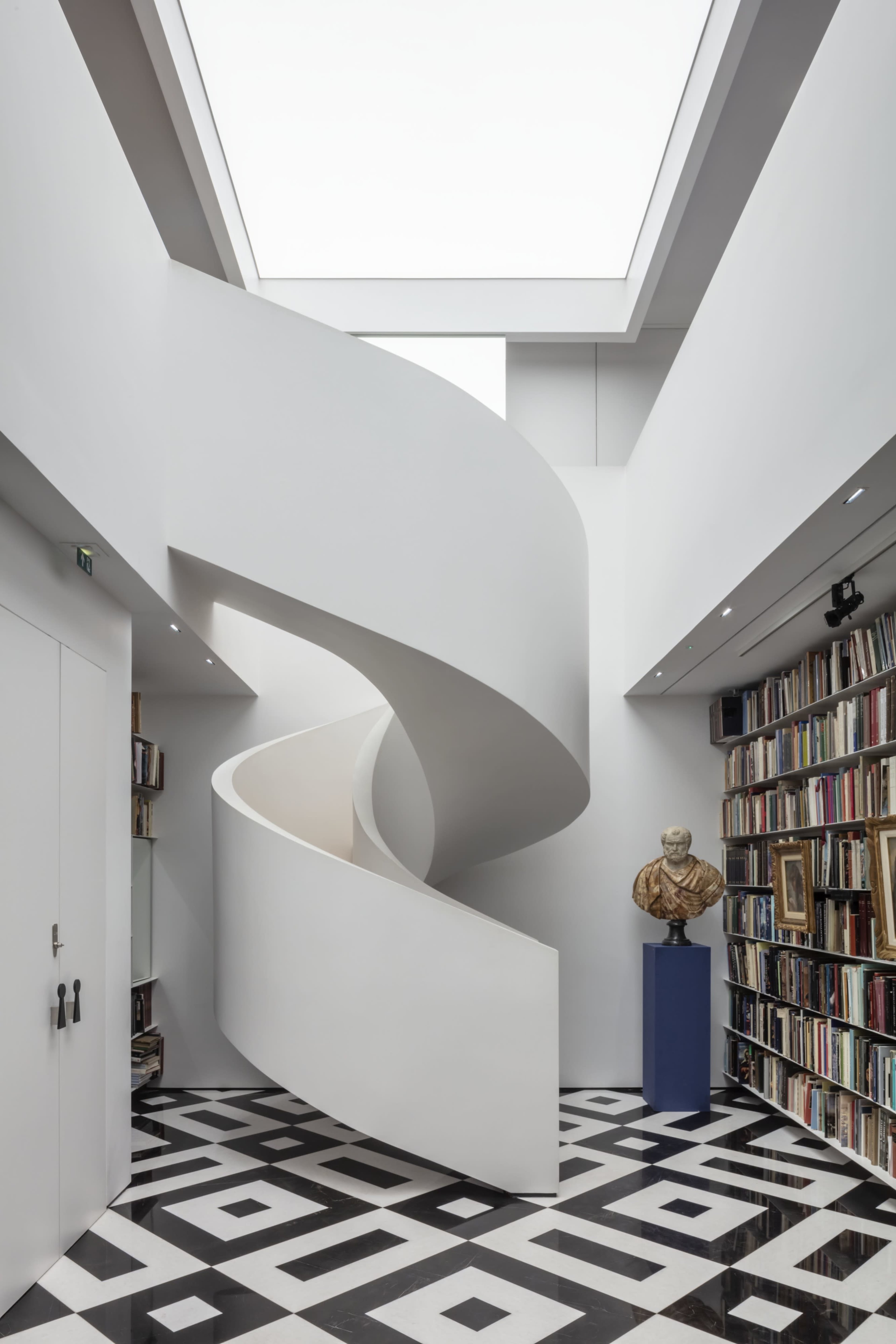 A modern interior with a curving white staircase at the center, surrounded by tall shelves filled with books and a bust sculpture on a pedestal.