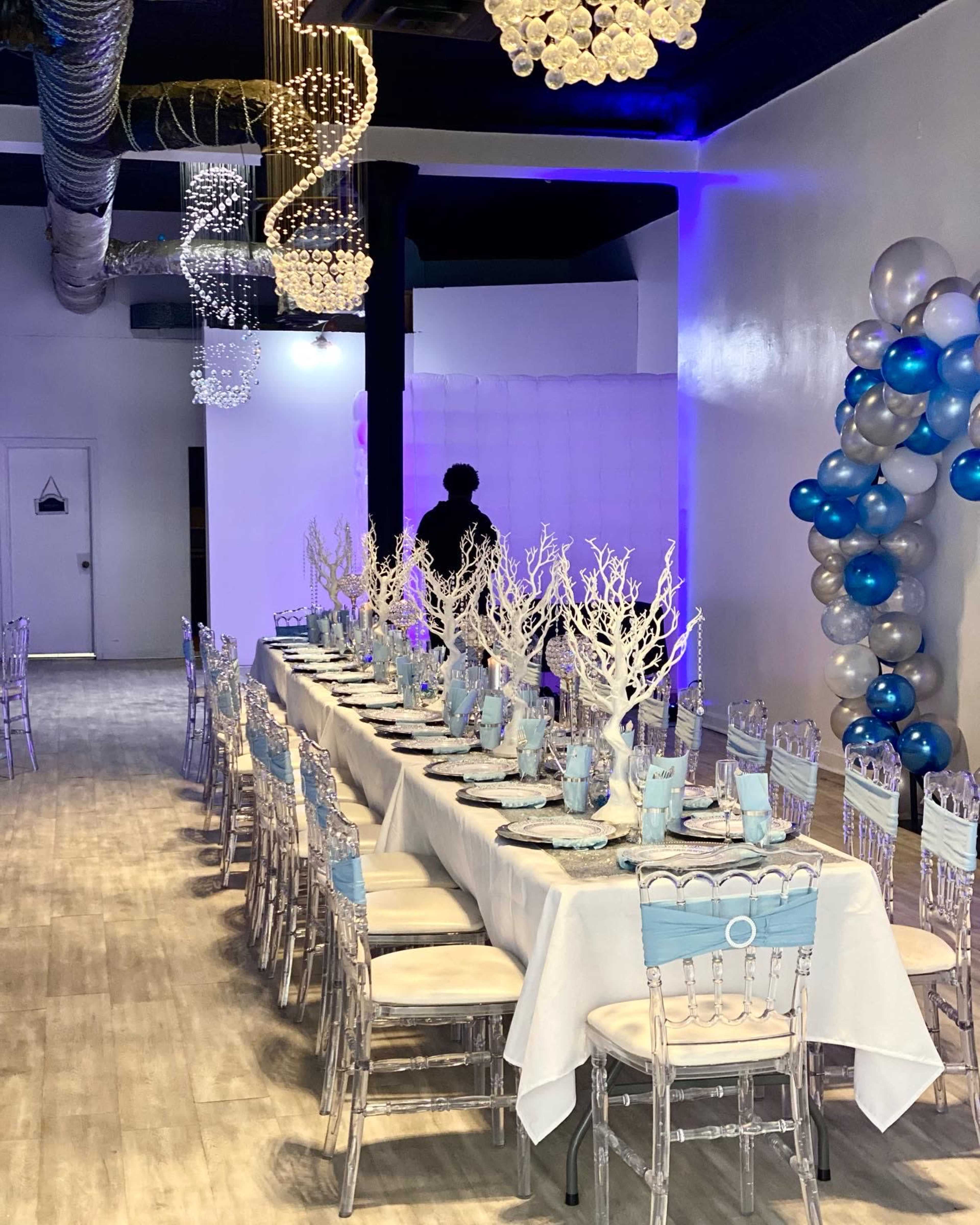 A beautifully arranged dining area with a long table set for an event, featuring clear chairs and decorative centerpieces of white branches, along with blue and silver balloons.
