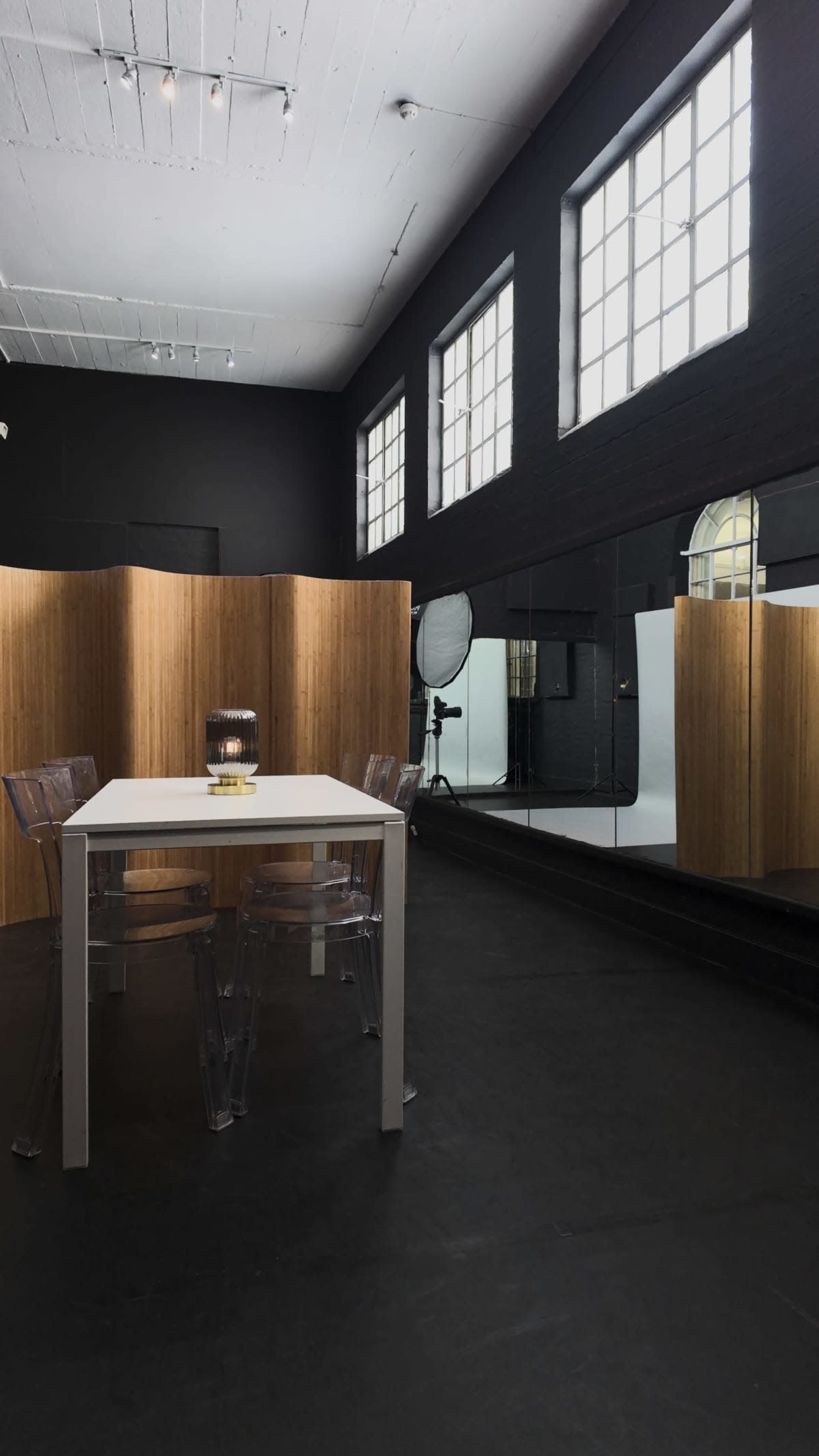 The image shows a modern studio space featuring a white dining table with transparent chairs, wooden partitions, and large windows that allow natural light to enter.