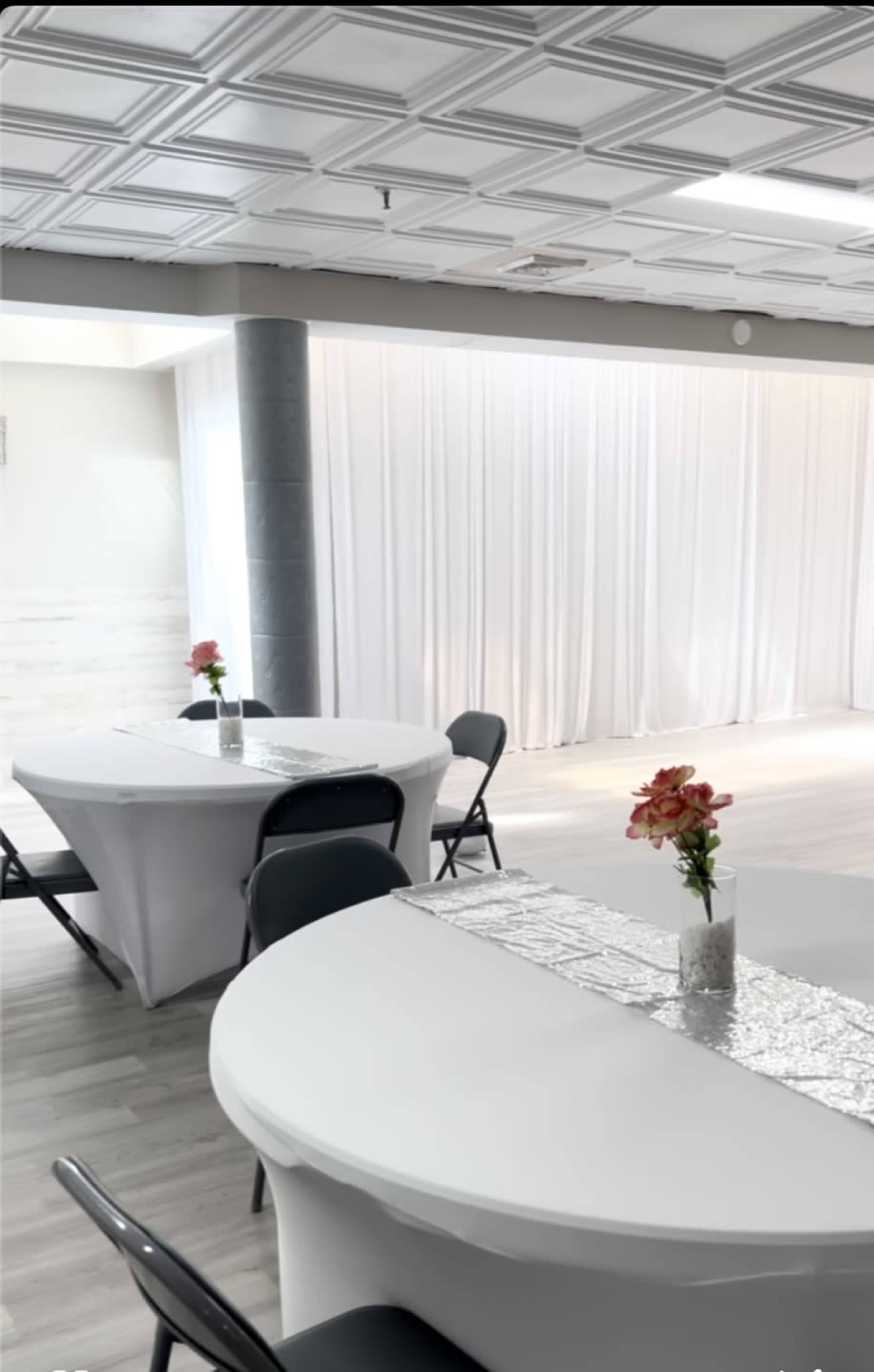 The image shows a bright event space with two round tables covered in white tablecloths, each adorned with a small vase of flowers, set against a backdrop of white drapery.