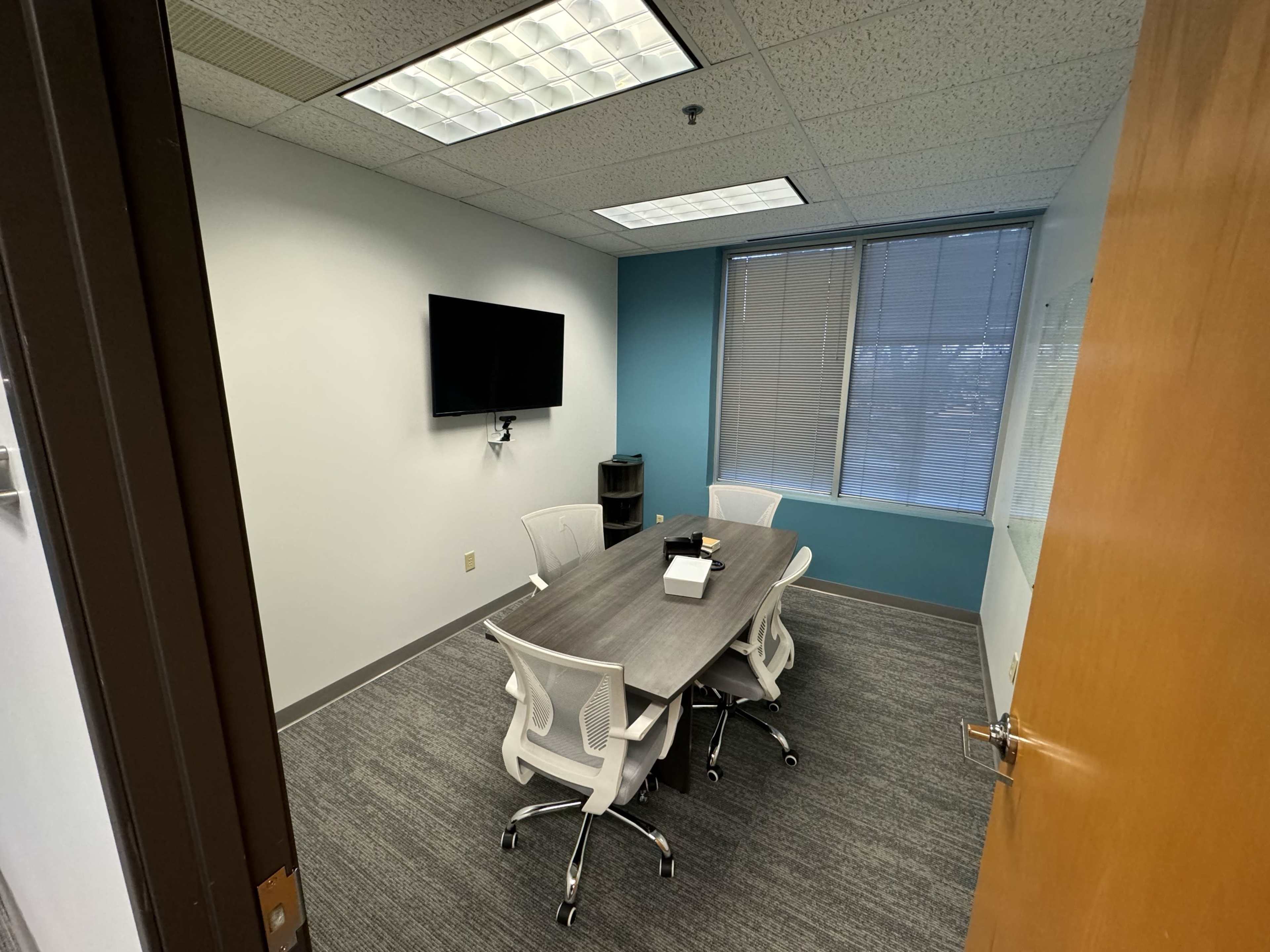 The image shows a small conference room with a rectangular table, four white chairs, a wall-mounted television, and large windows providing natural light.