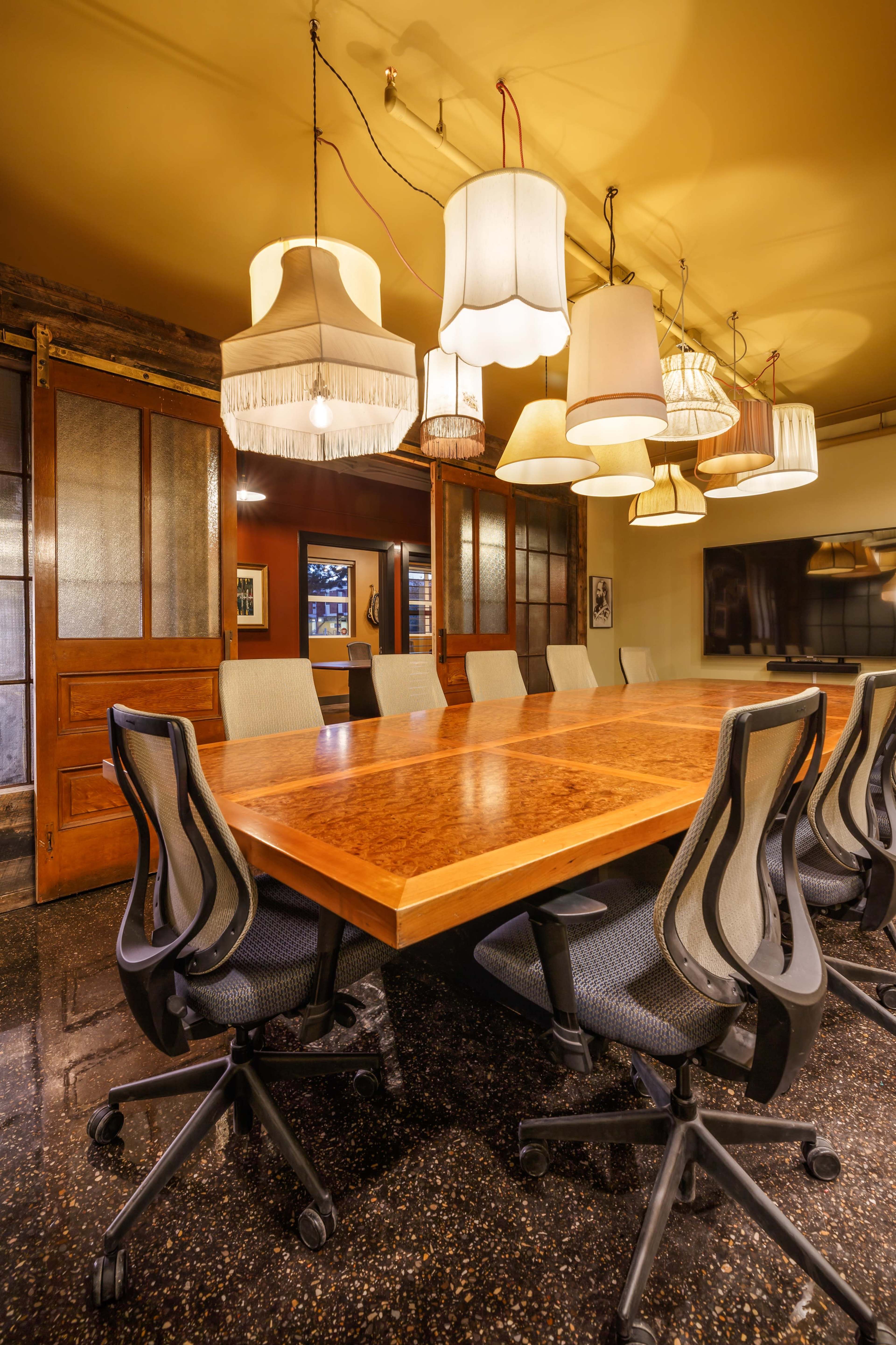 A long wooden conference table surrounded by ergonomic chairs is illuminated by several pendant lamps hanging from the ceiling.