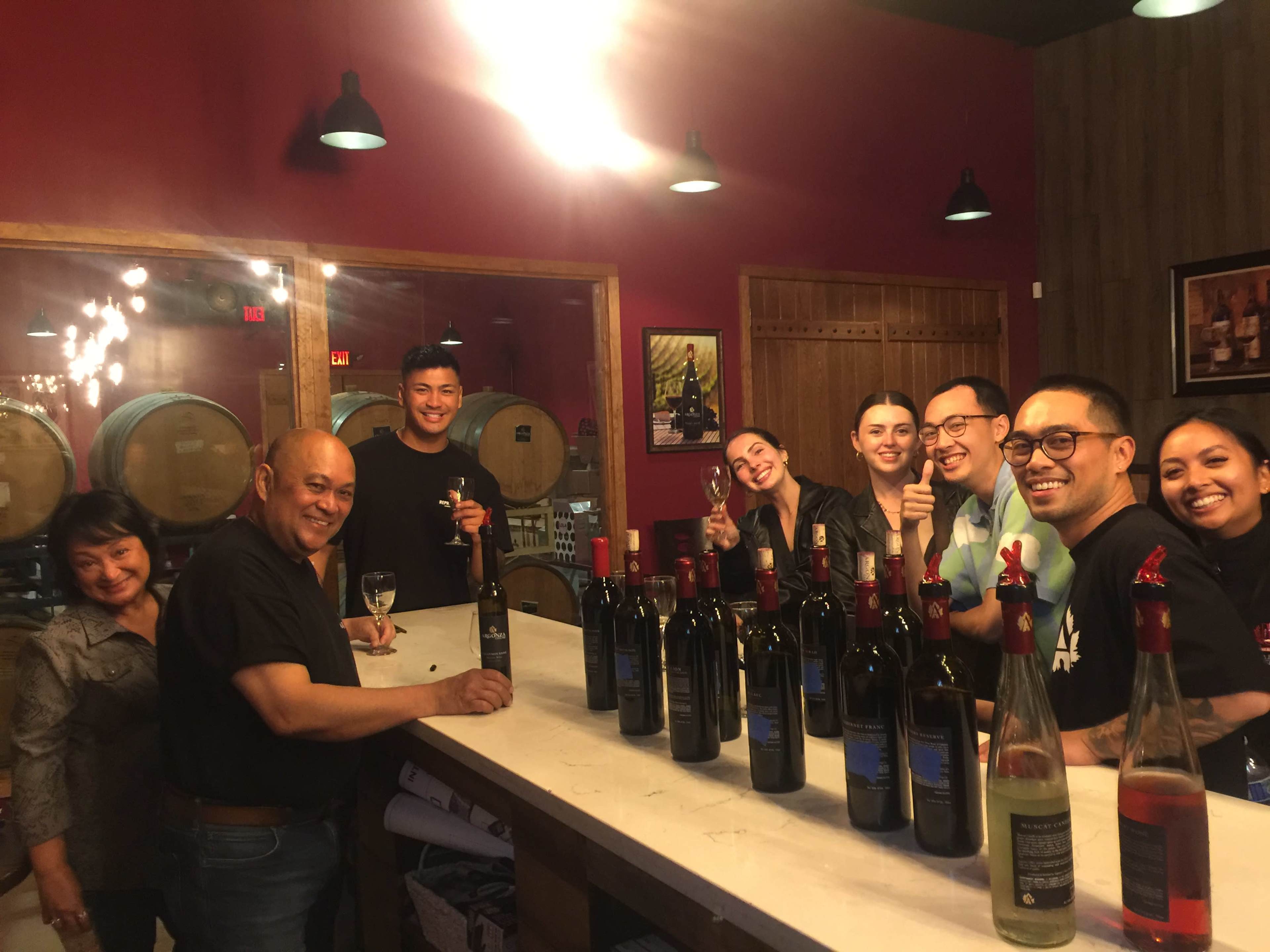 A group of people is gathered at a bar surrounded by wine bottles, smiling and posing for the camera.
