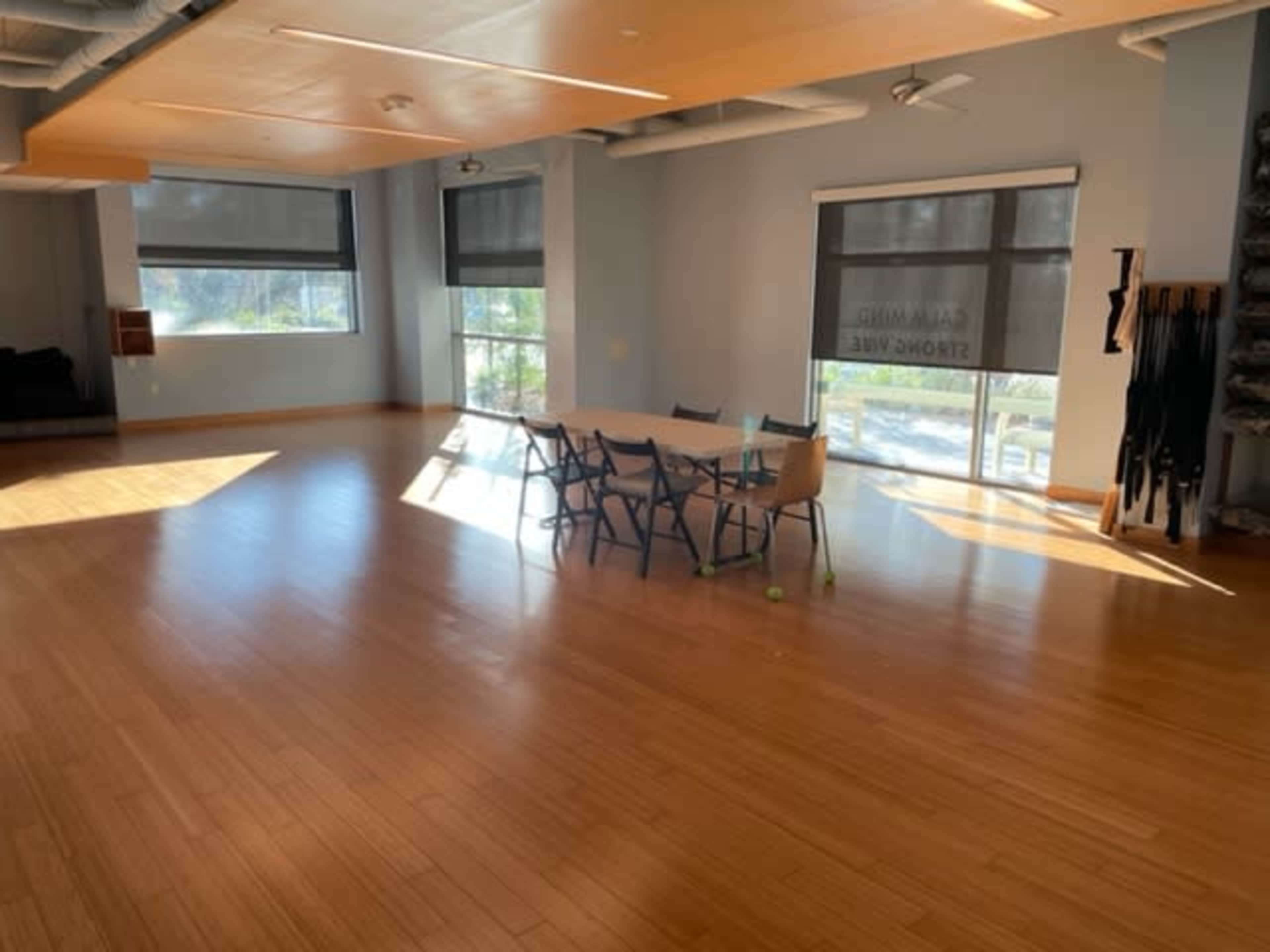 A spacious room with wooden flooring features a central table surrounded by chairs, large windows with blinds, and minimal furnishings along the walls.
