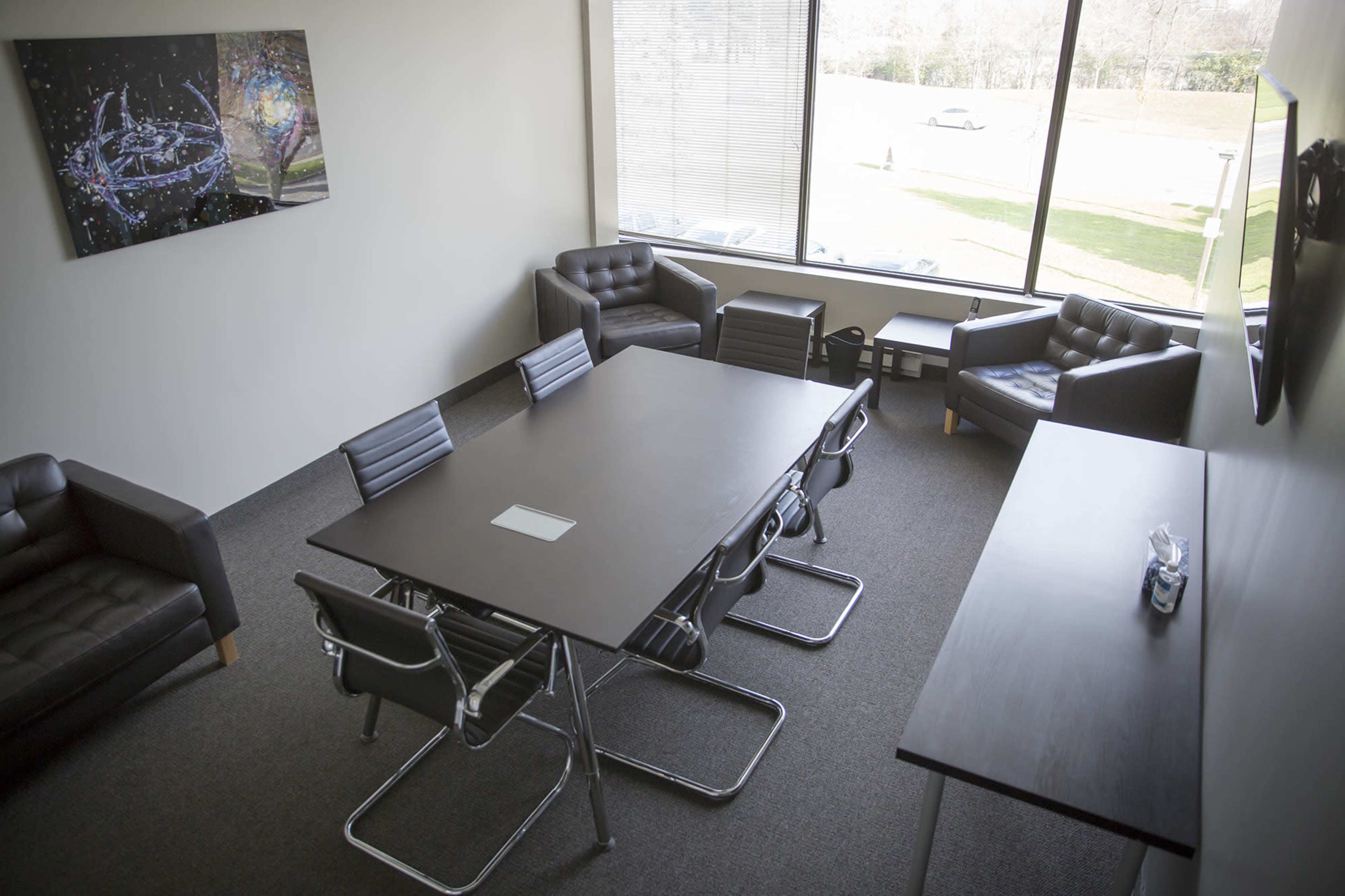 A conference room features a large table surrounded by chairs, with two armchairs in the corner and a large window overlooking an outdoor area.