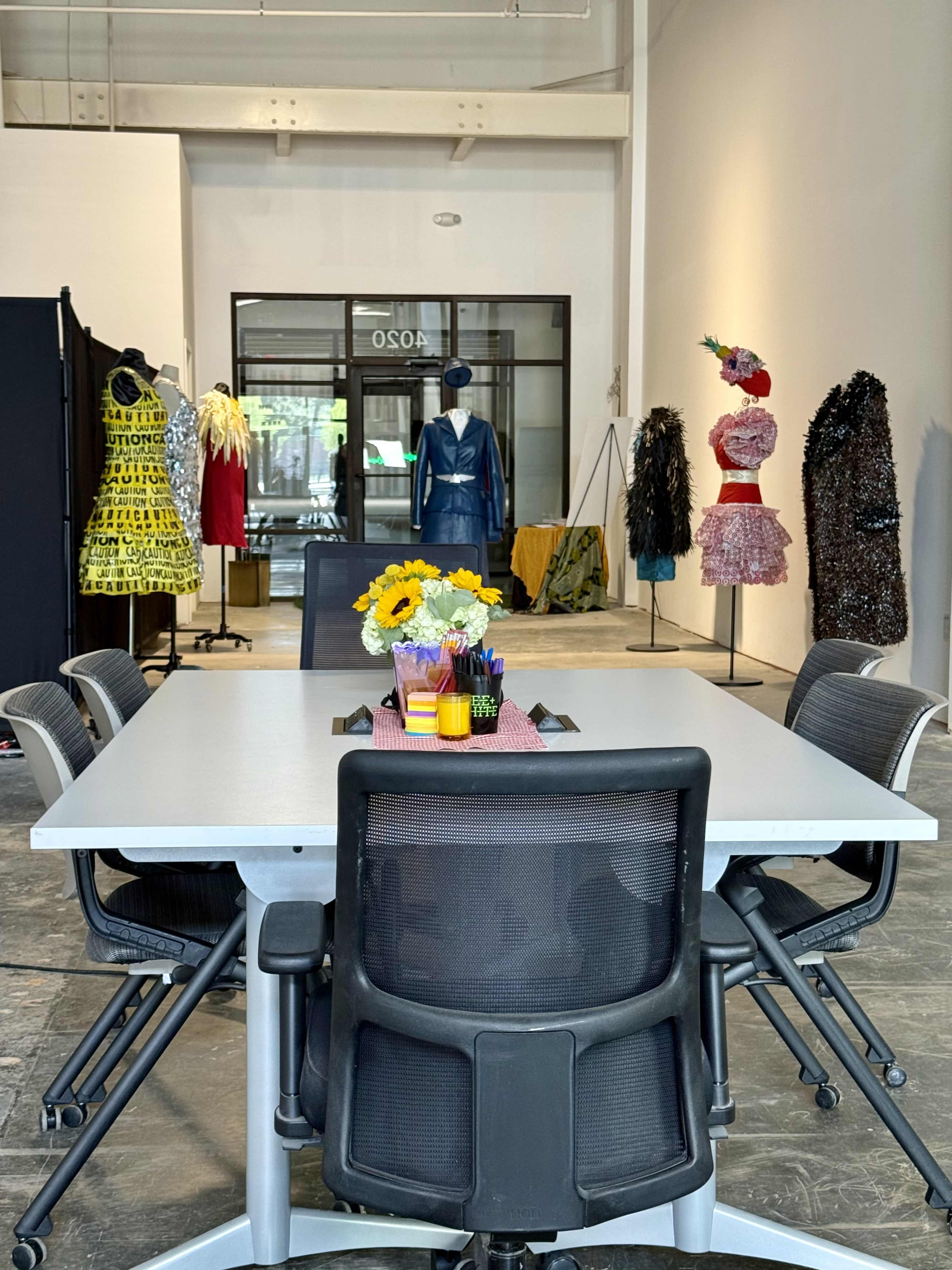 A modern meeting room features a large table surrounded by black chairs, with colorful dresses displayed on mannequins in the background.