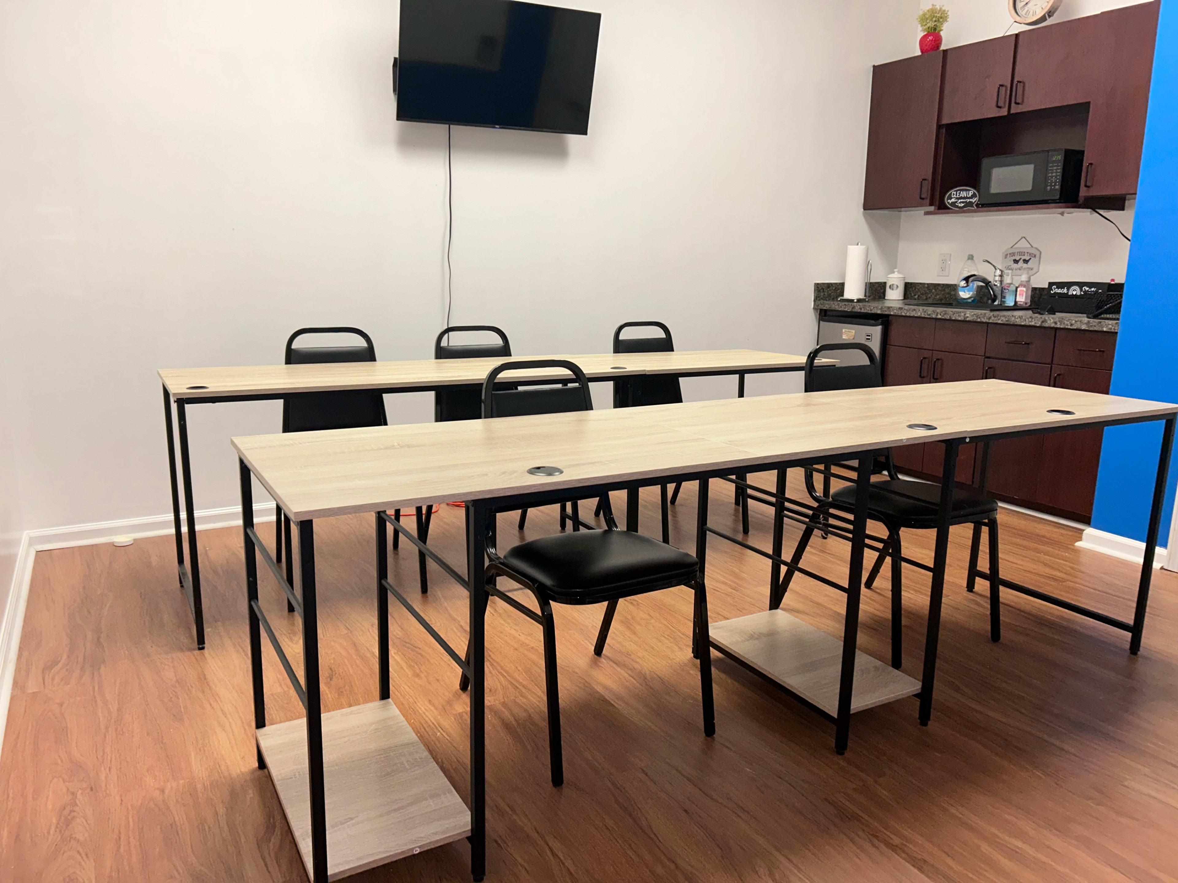 The image shows a small meeting room with two long tables arranged in a U-shape, black chairs, and a television mounted on the wall.