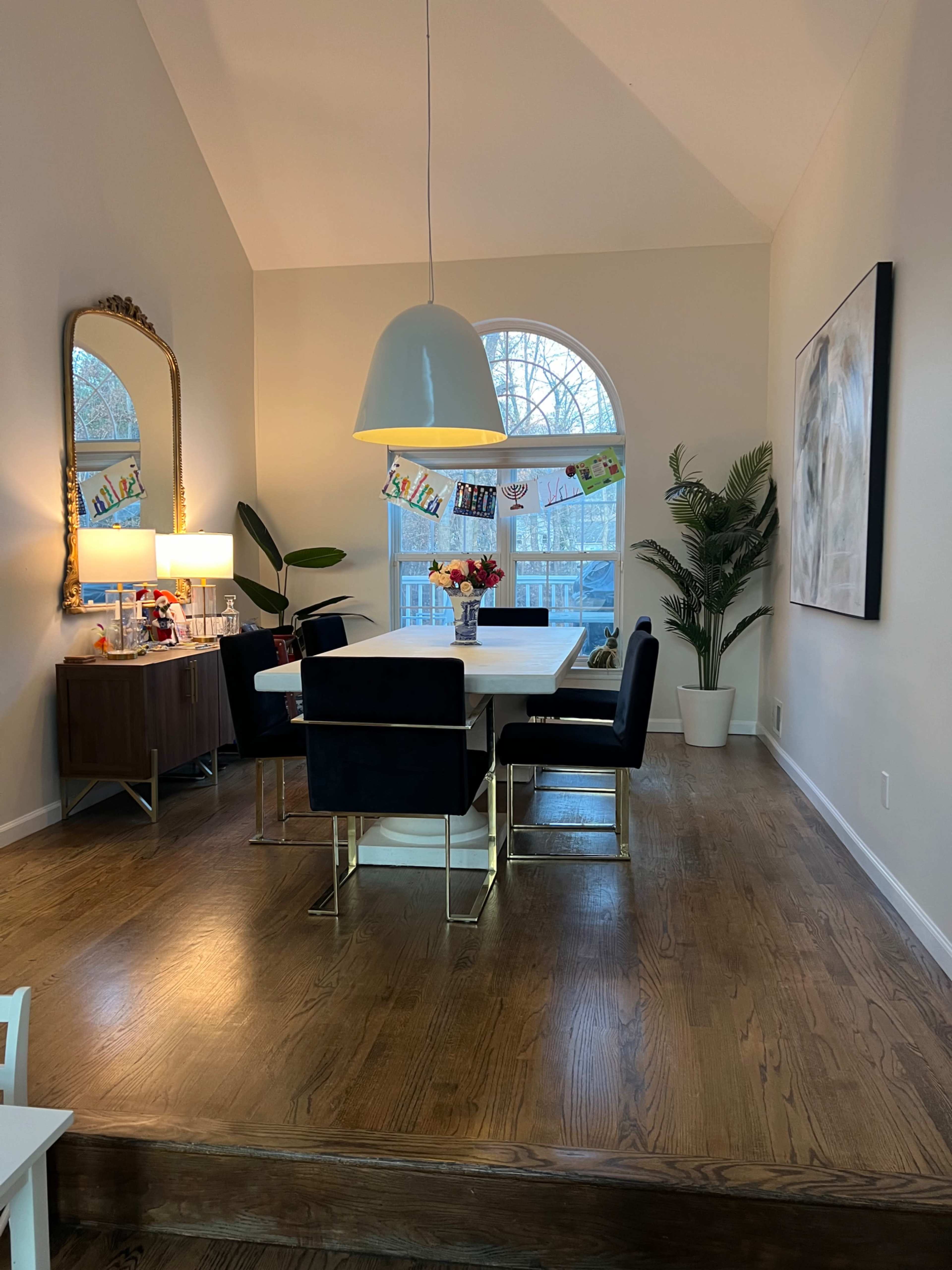A dining area features a large white table surrounded by black chairs, with a decorative mirror and a potted plant in the corner.