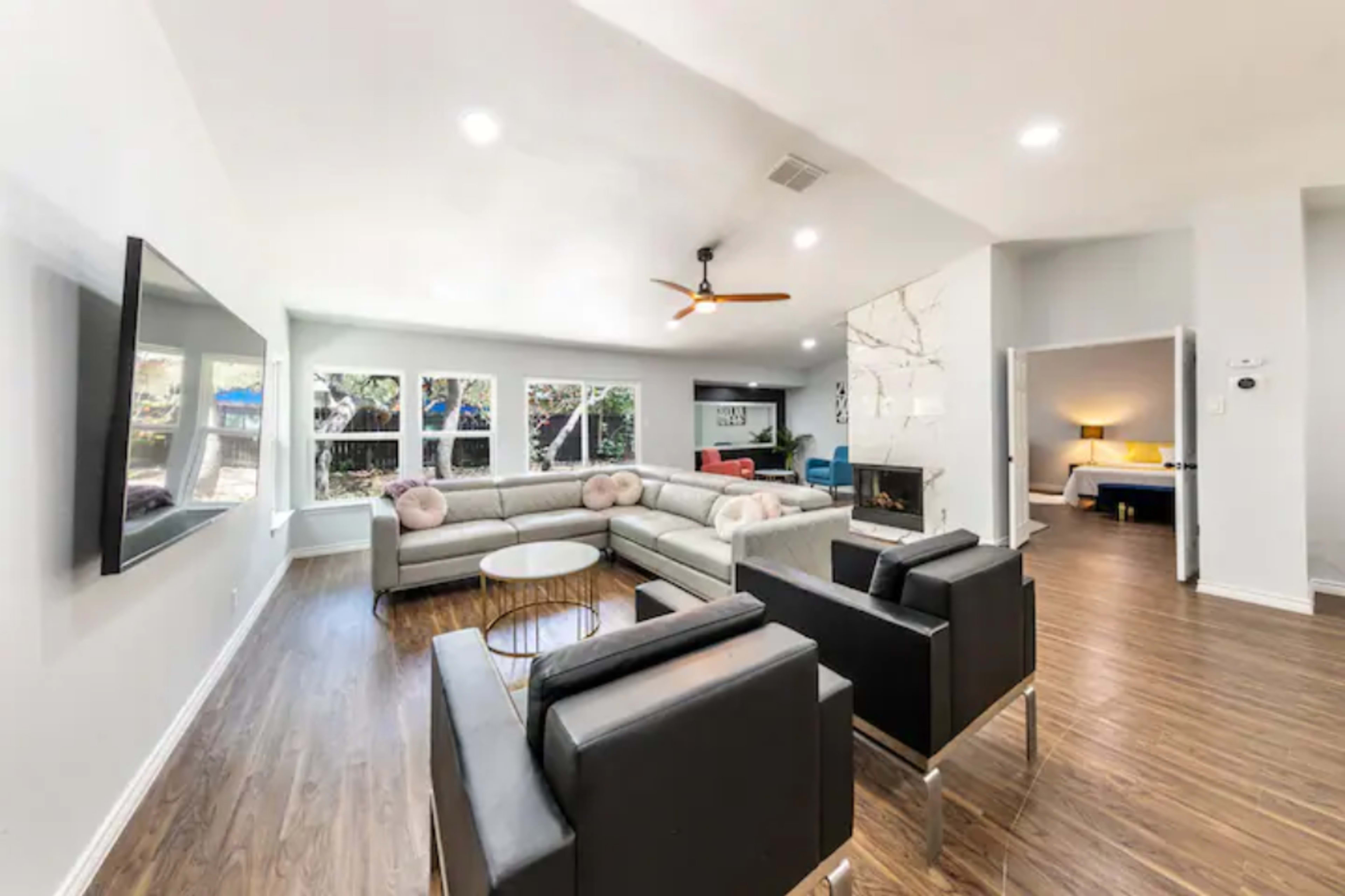 The image shows a modern living room featuring a sectional sofa, two black armchairs, a round coffee table, and large windows letting in natural light.