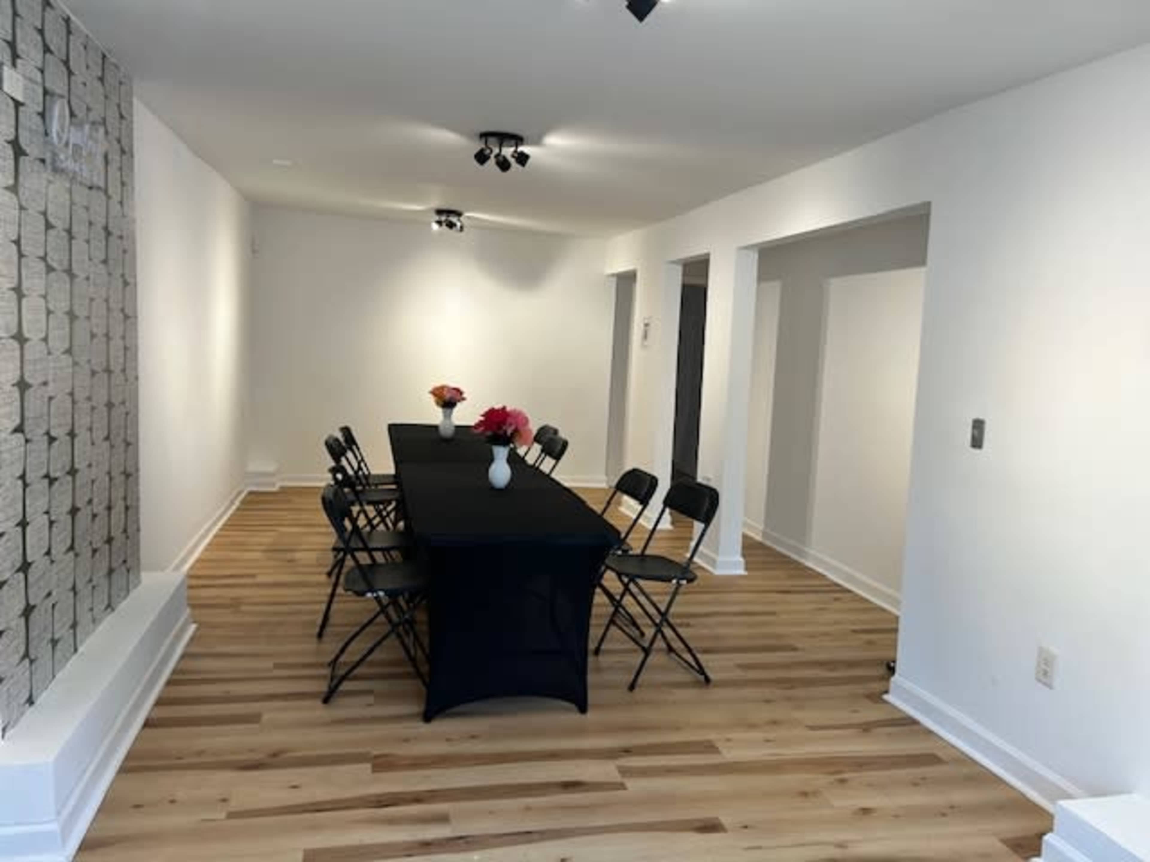 A simple, well-lit room features a long black table surrounded by folding chairs, with a vase of flowers at the center and a patterned wall in the background.