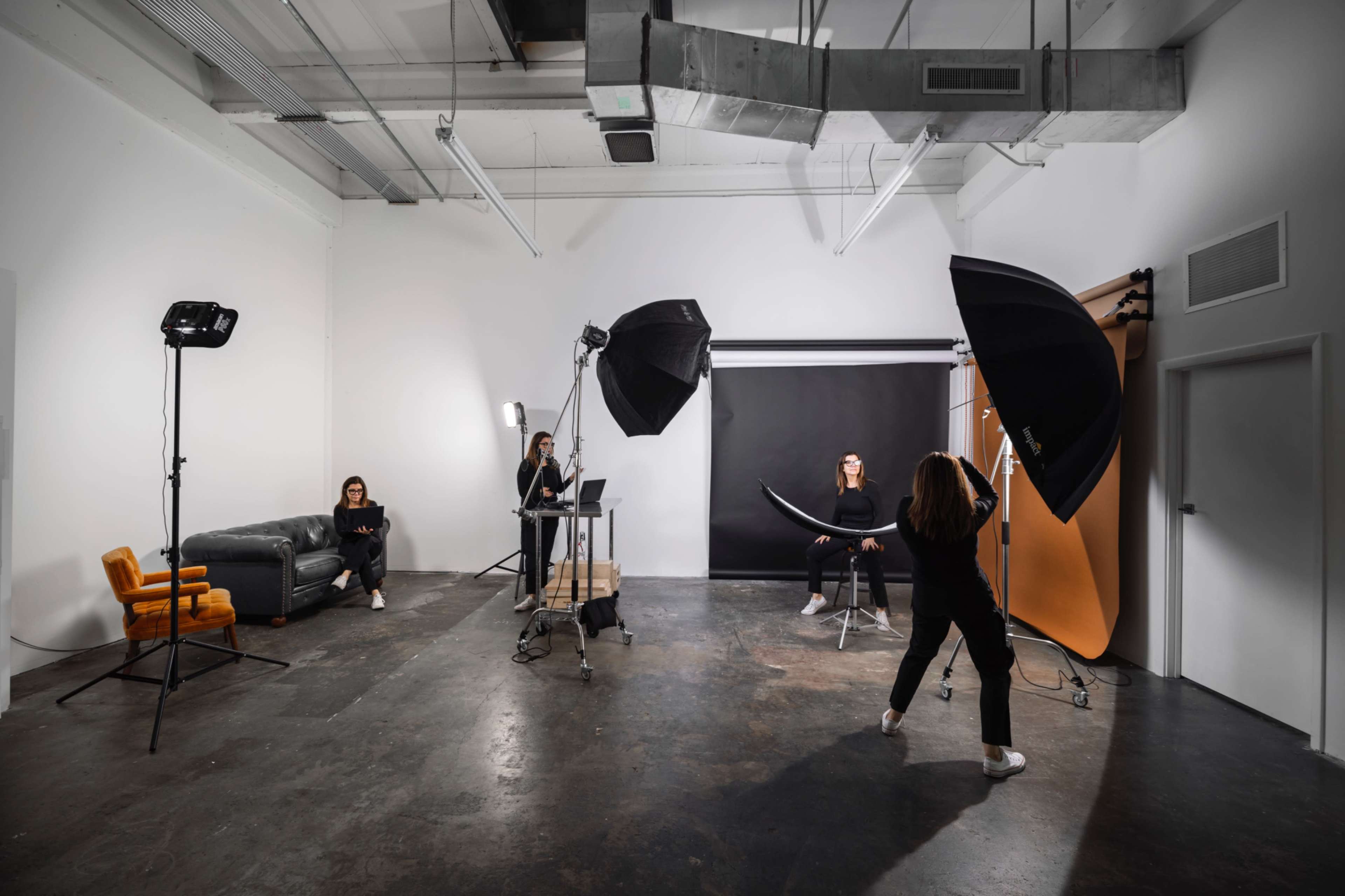 A photography studio is set up with multiple lighting setups, a model sitting on a stool, and two photographers working at a computer and adjusting equipment.