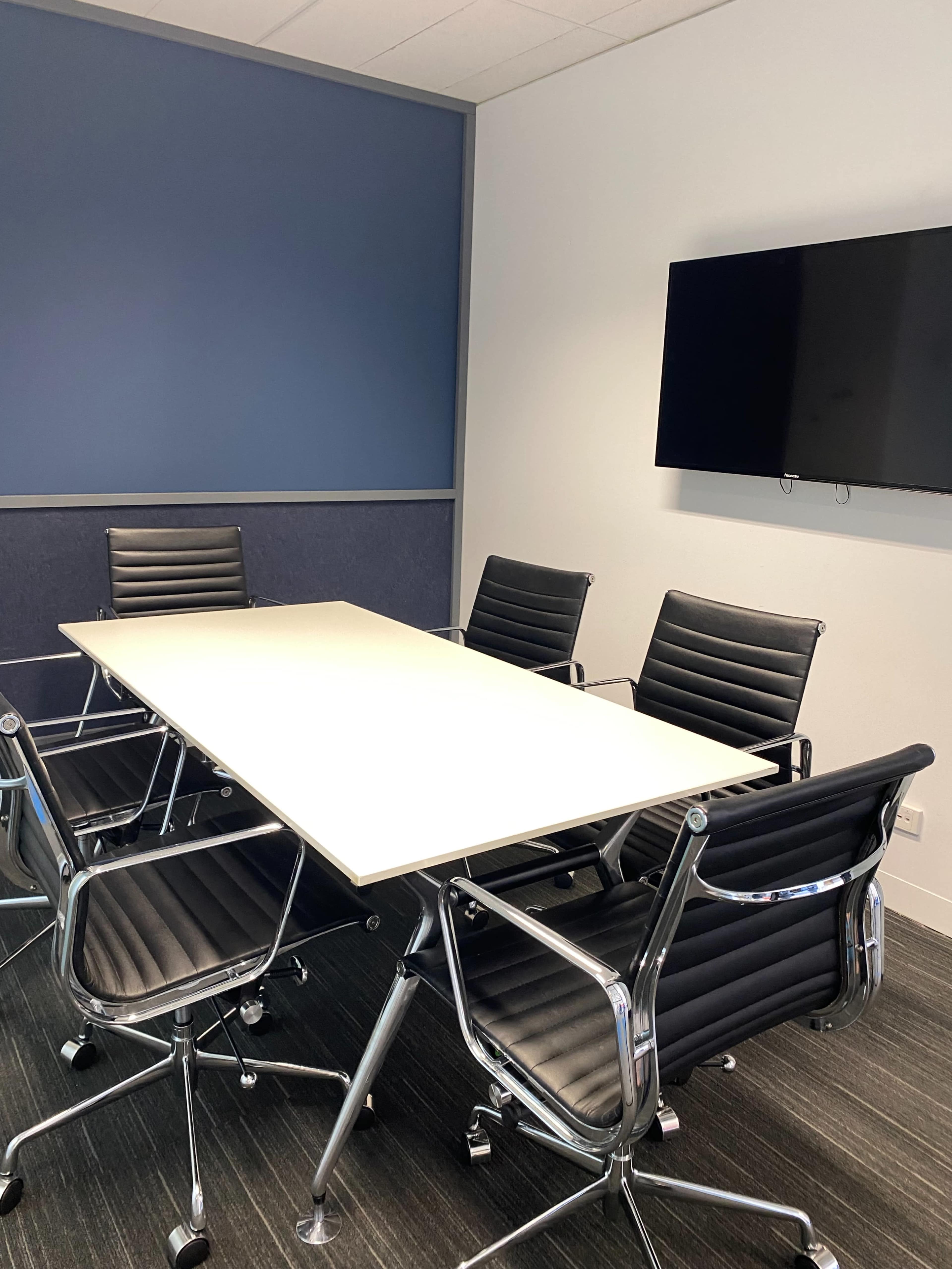 A conference room features a rectangular table surrounded by six black chairs and a wall-mounted television.