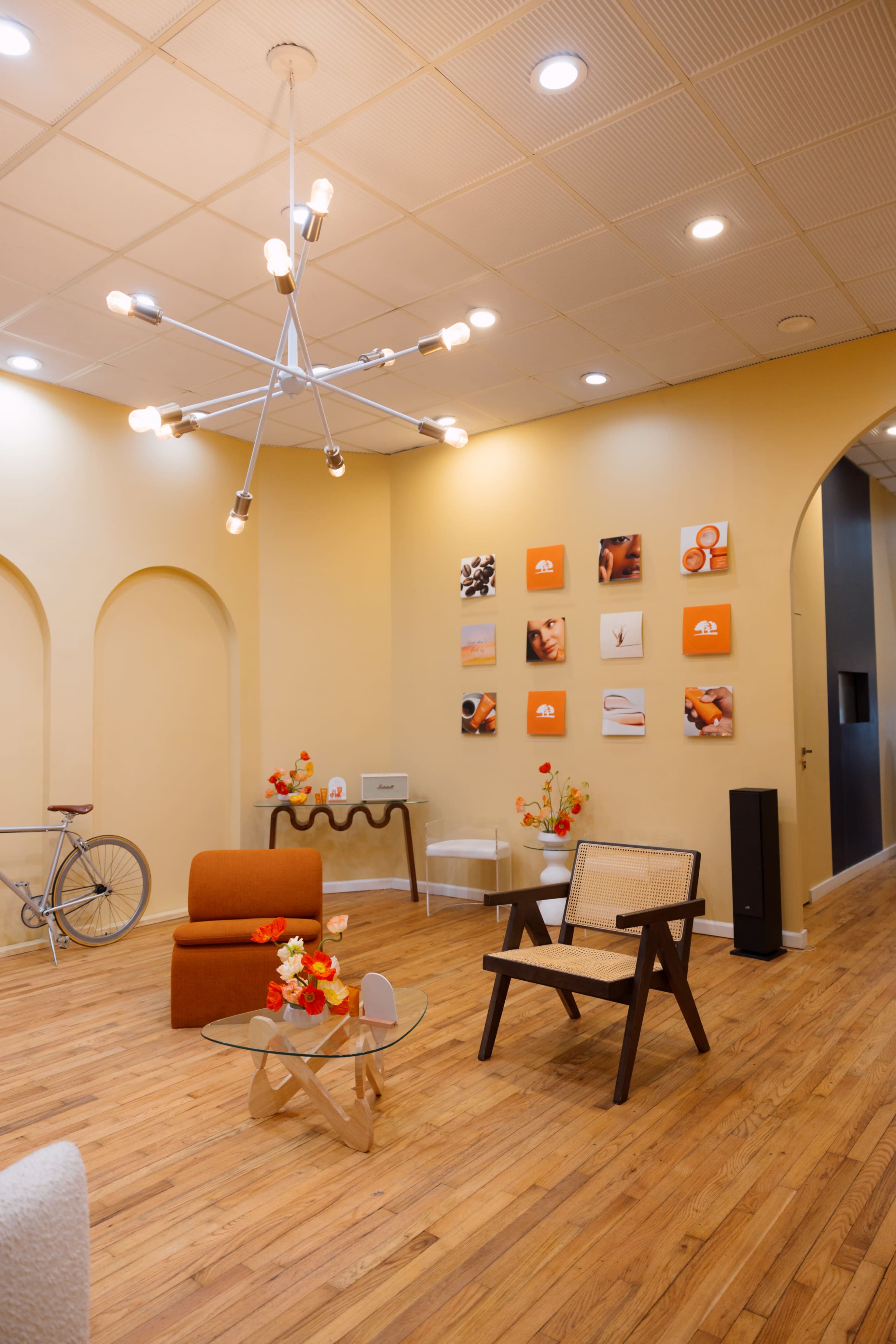 The image shows a modern interior space with a bicycle leaning against the wall, orange and white decorative elements, and various seating options arranged around a glass coffee table.