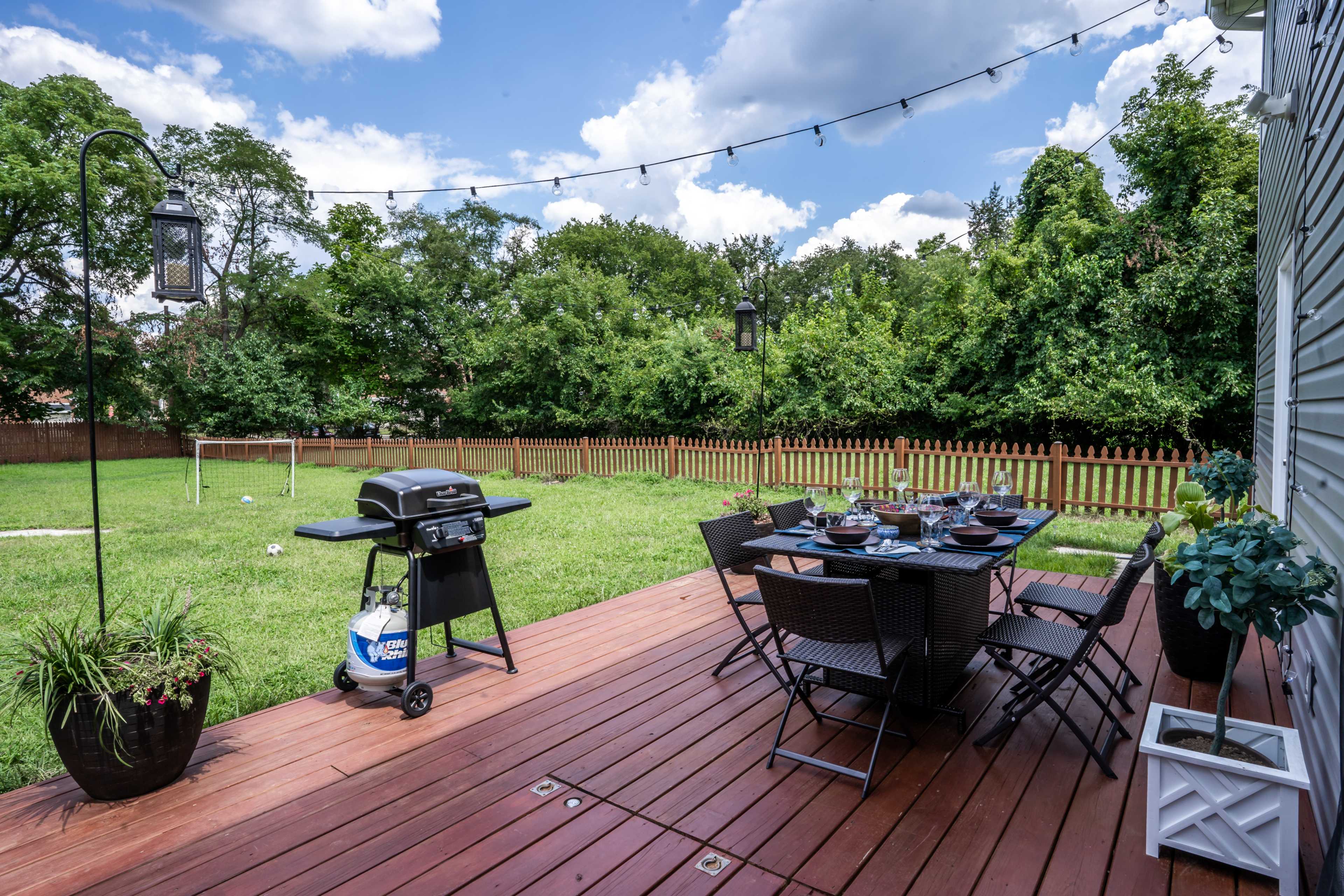 A wooden deck features a dining table set with dishes and a grill, surrounded by a green lawn and trees in the background.