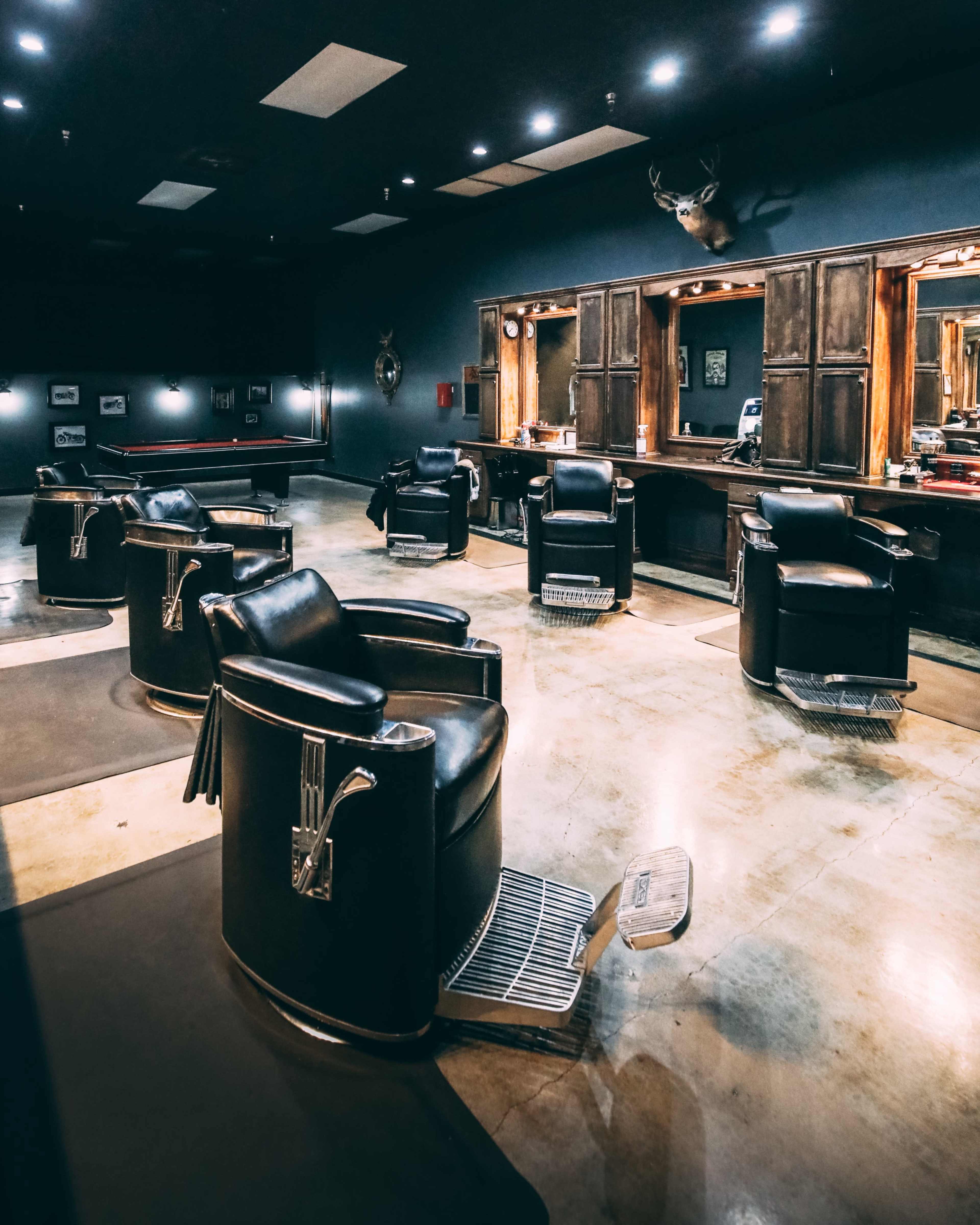 A spacious barber shop with several black leather barber chairs arranged in front of wooden mirrors and a contrasting dark wall.