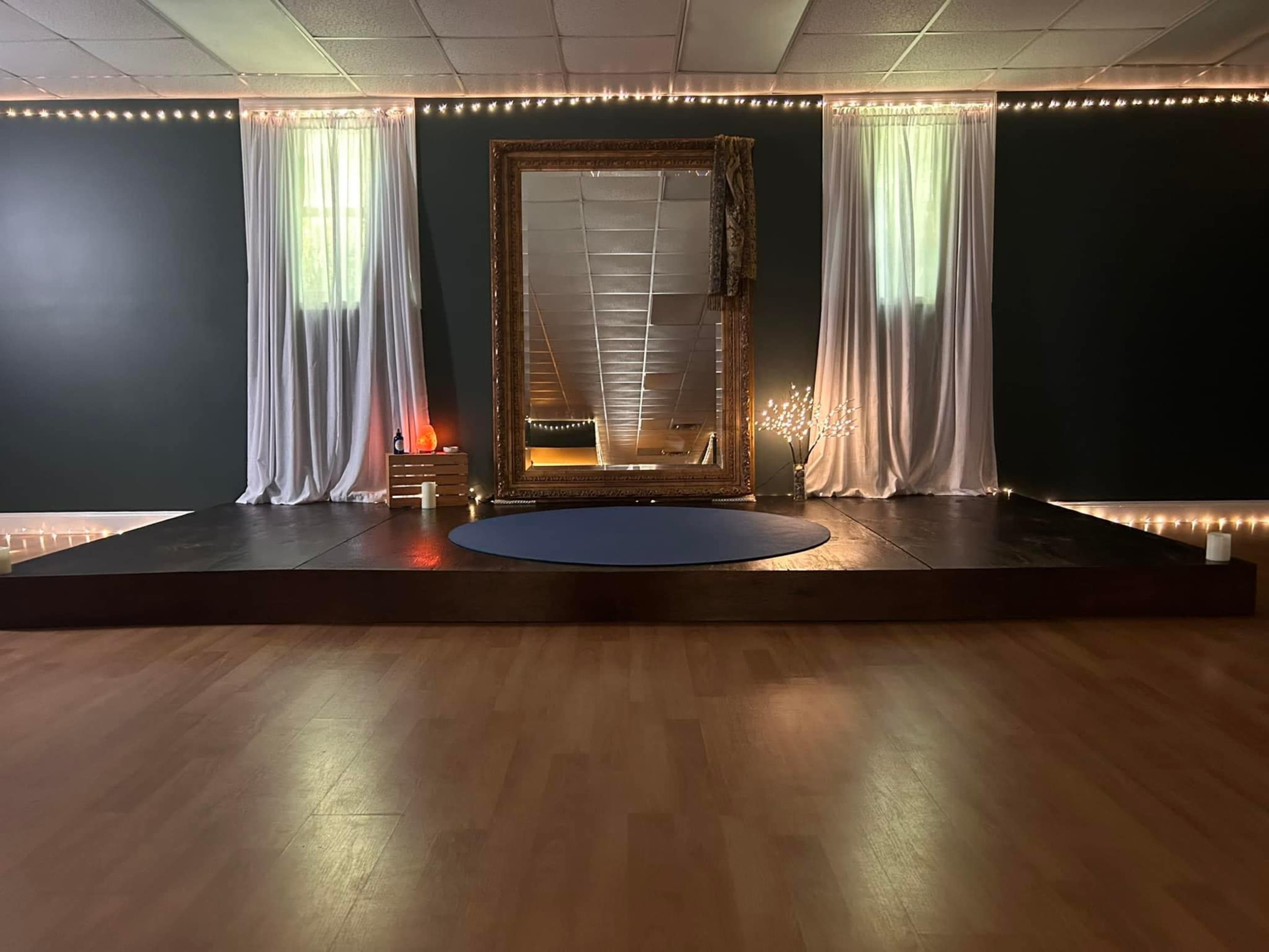 A spacious room with a large ornate mirror framed in gold, flanked by sheer white drapes, and dim lighting complemented by a circular blue mat on a wooden stage.
