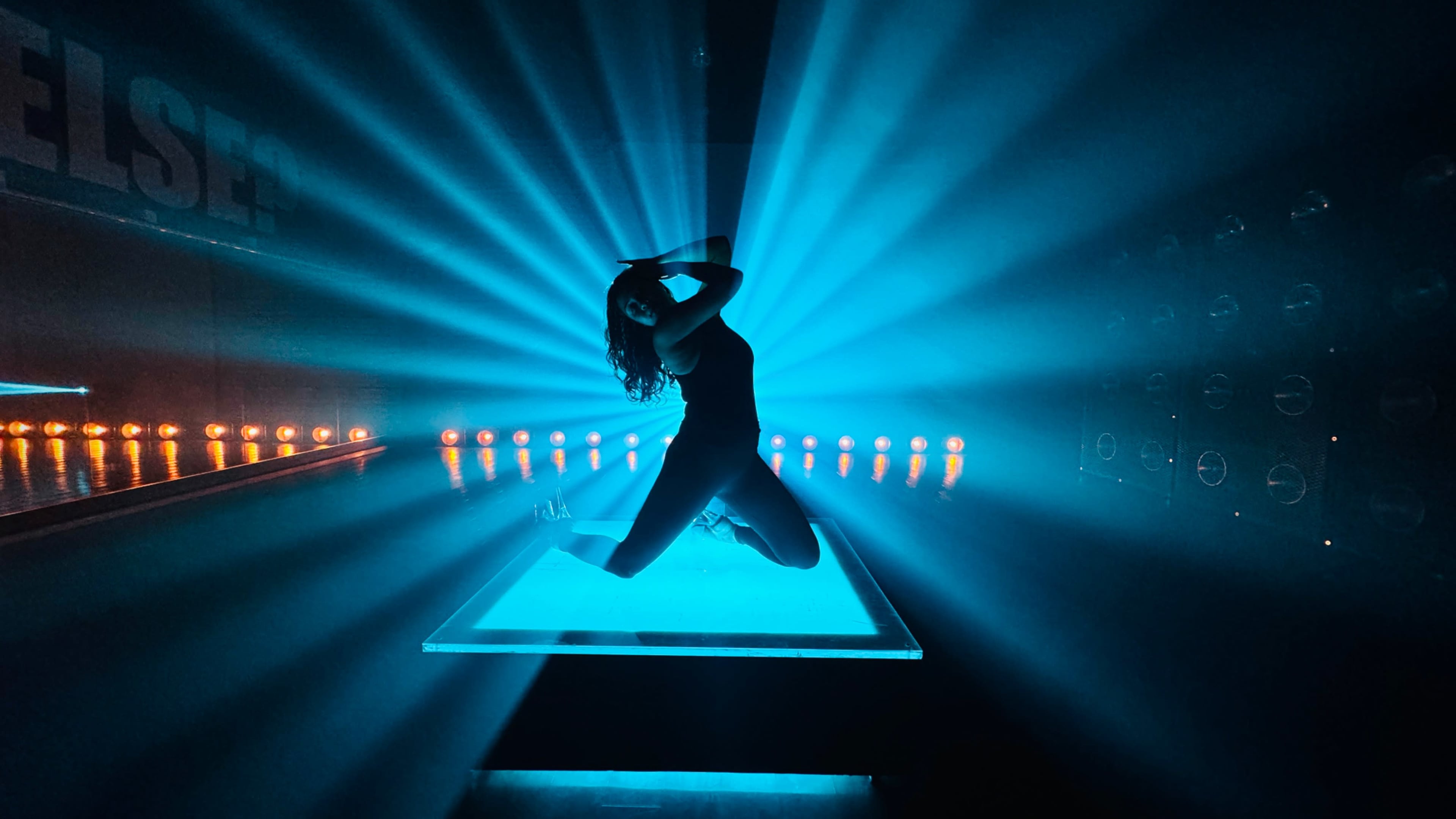 A silhouetted dancer poses dramatically on a illuminated platform with beams of blue light radiating from behind.