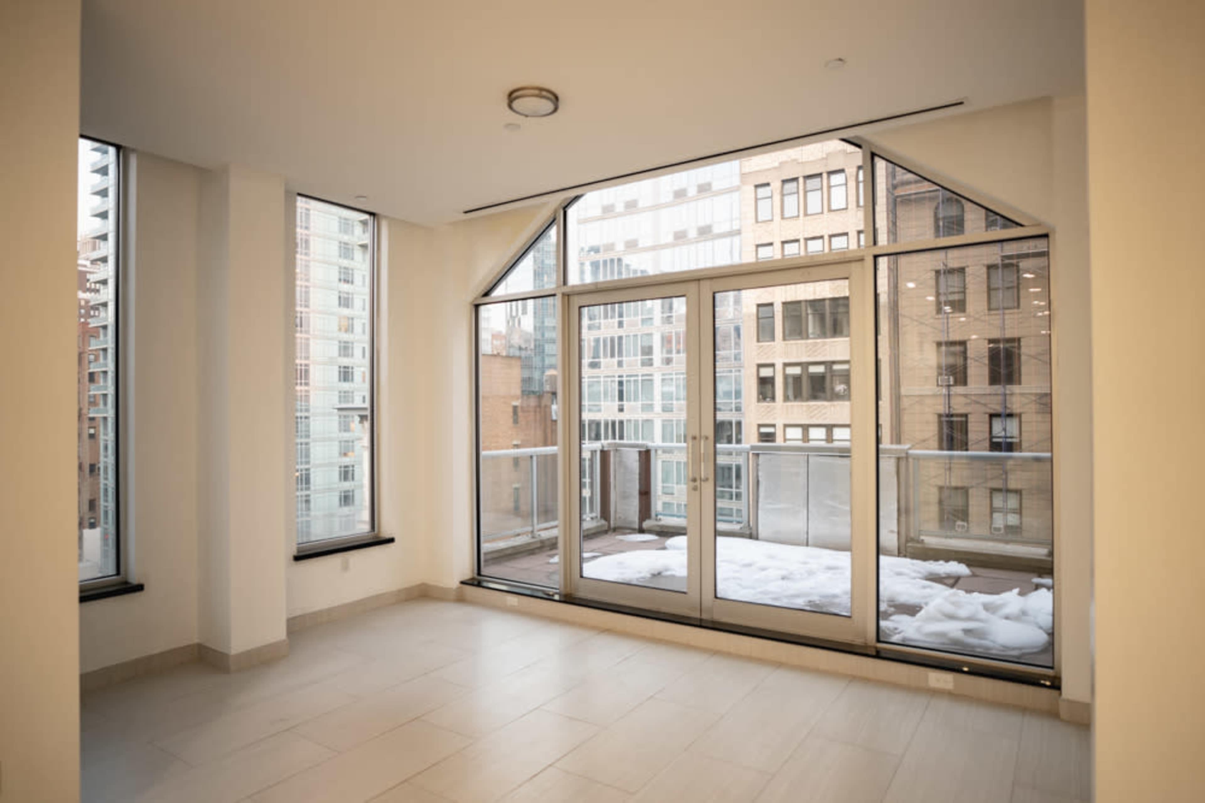 The image shows a vacant room with large windows overlooking a cityscape, featuring a balcony with some snow on it.