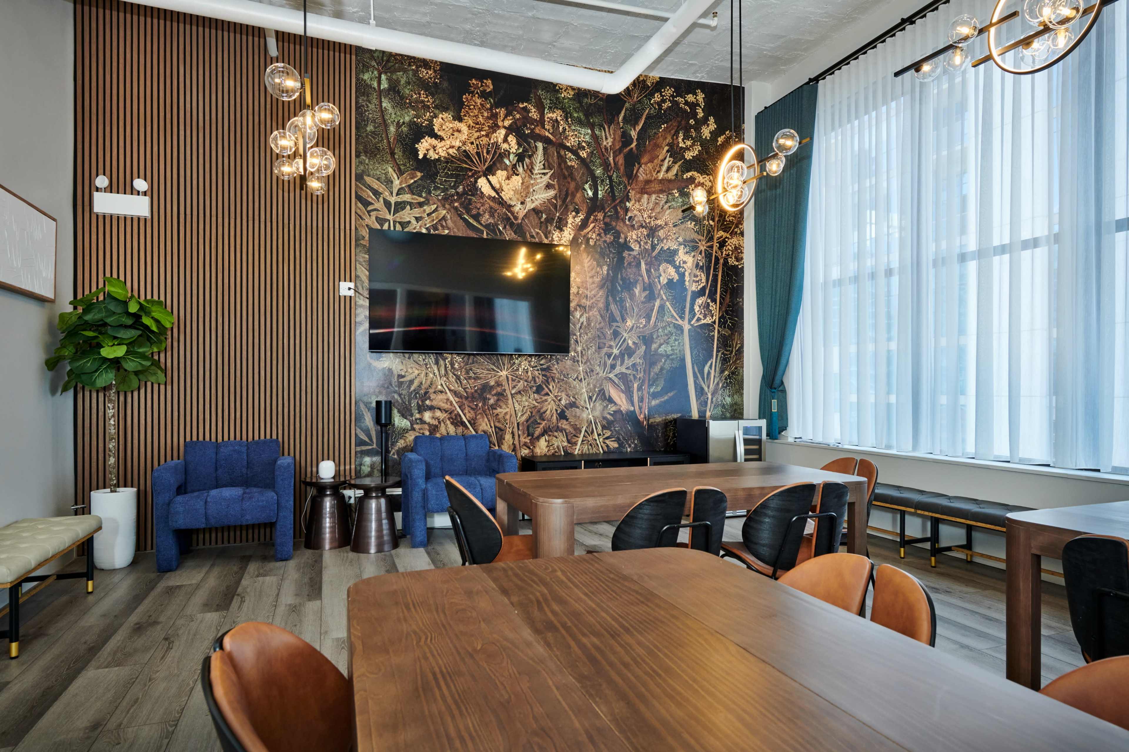 A modern meeting room with wooden flooring, a large wall-mounted TV, and seating areas featuring blue chairs and a decorative plant.