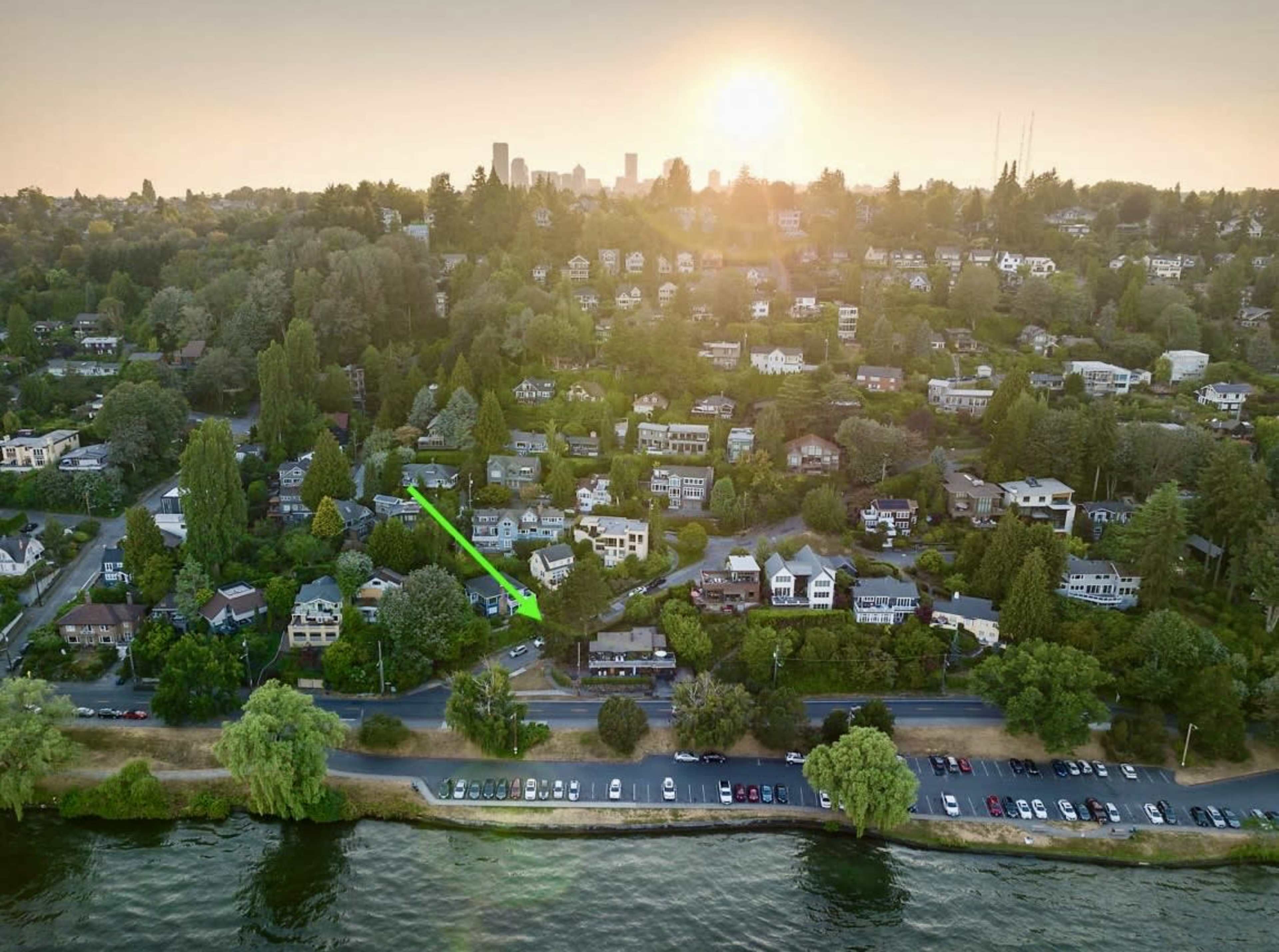 A waterfront view of a residential area with houses nestled among trees, and the skyline of a city visible in the background under a setting sun.