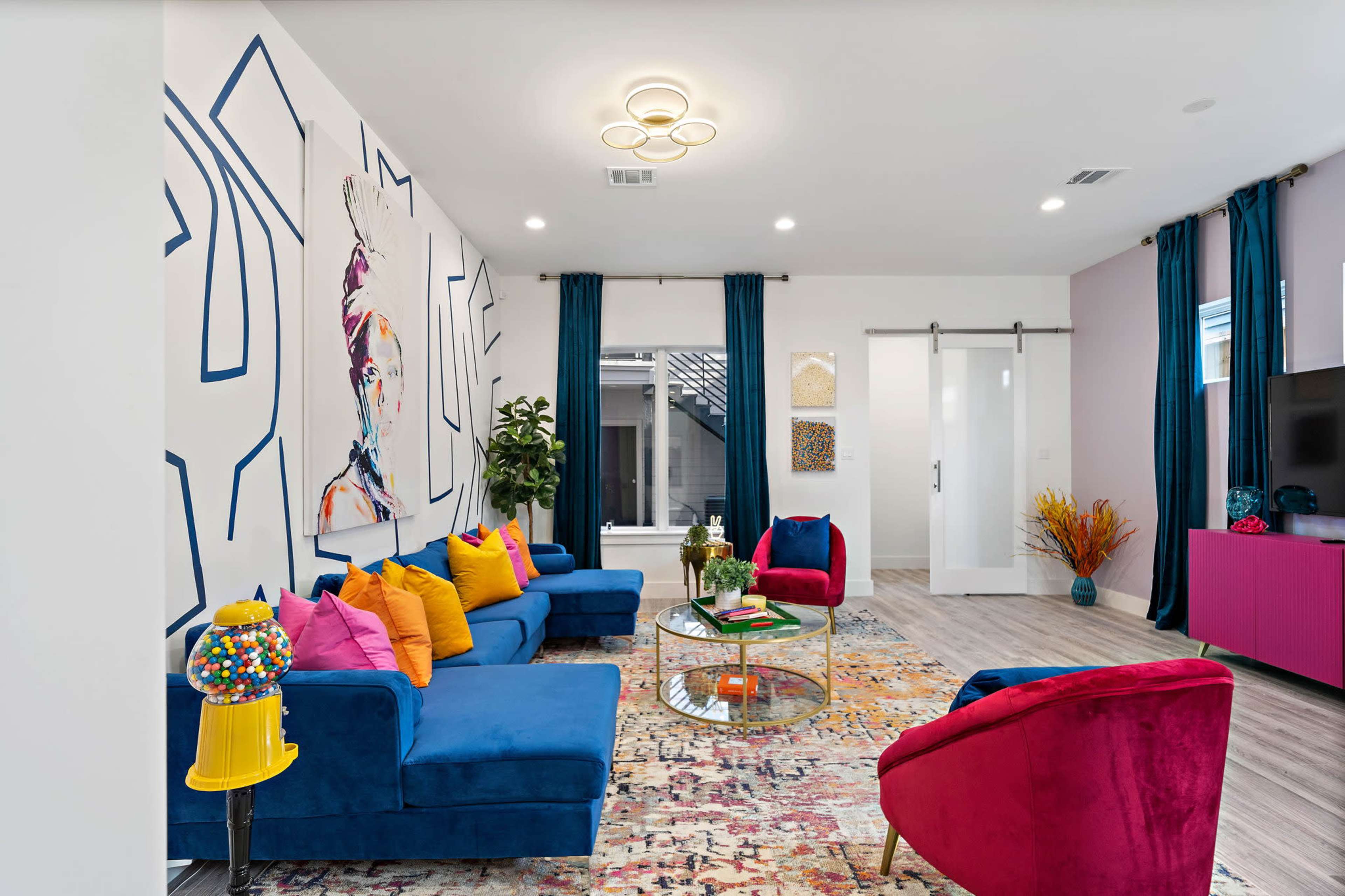 The room features a vibrant seating area with blue sofas, colorful pillows, and a round glass coffee table, surrounded by large artwork and decorative elements.