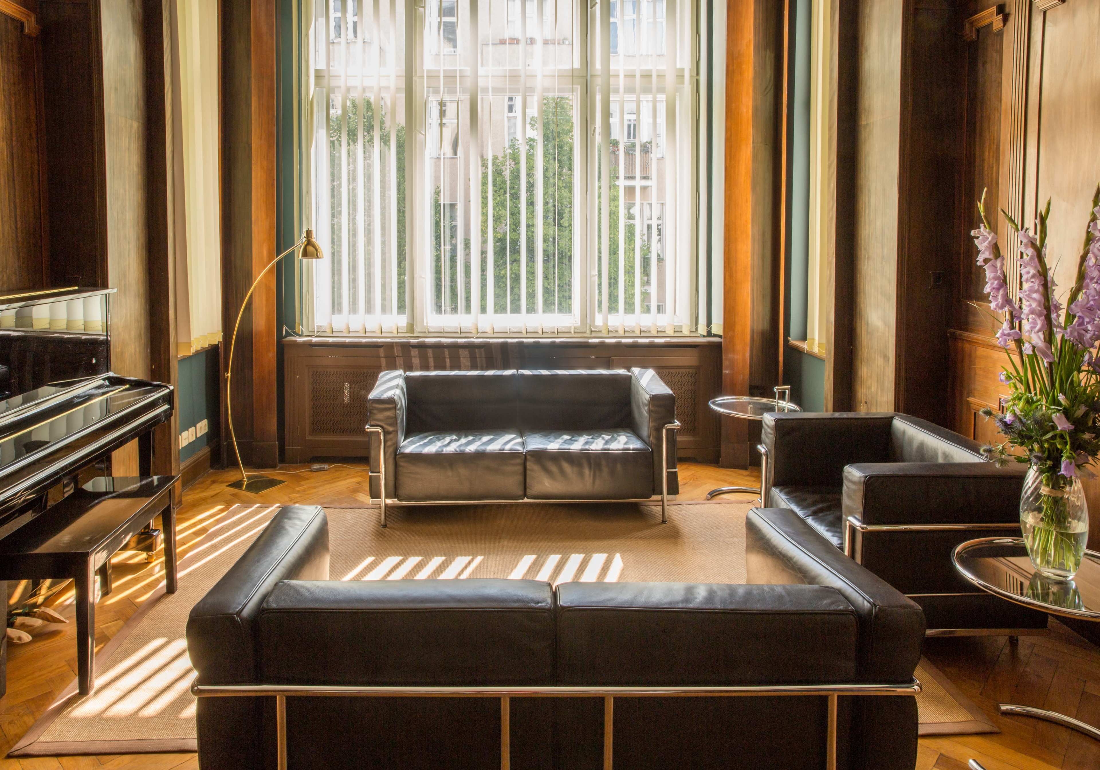 A spacious room with a black leather sofa and chairs arranged around a central area, illuminated by natural light from large windows with vertical blinds.