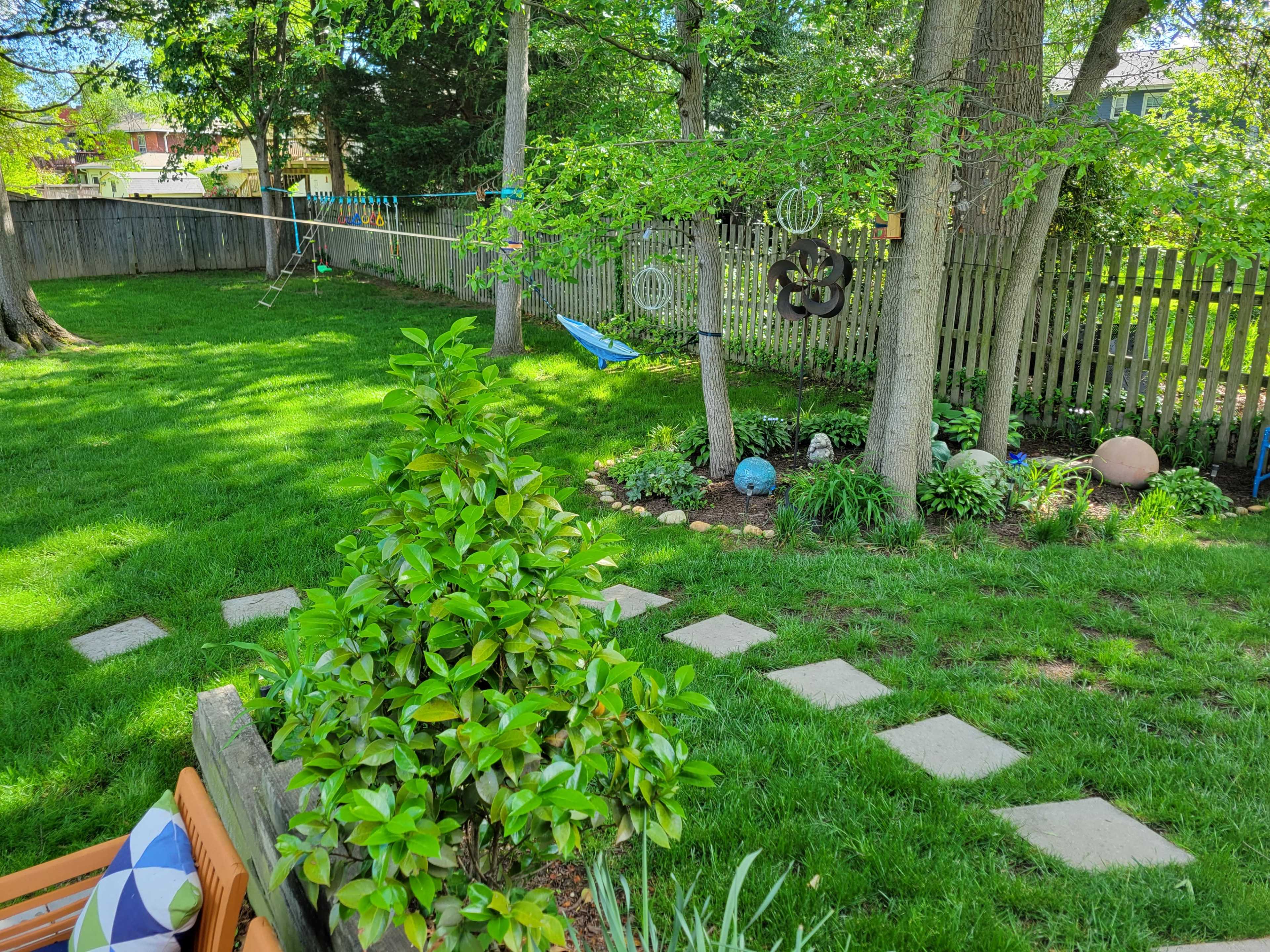 The image shows a backyard with green grass, a stone pathway, trees, a clothesline, and decorative objects among the foliage.