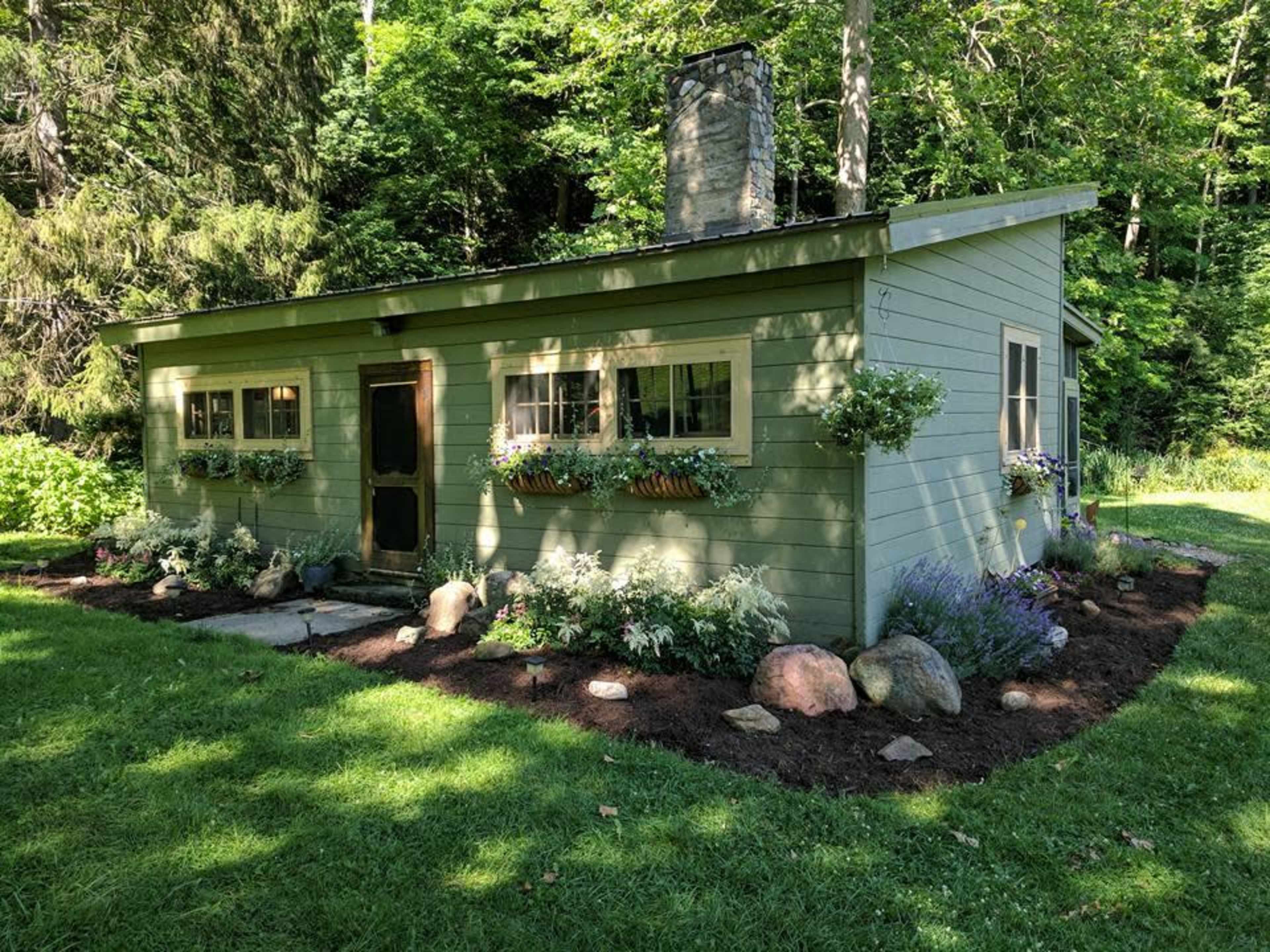 A small green cottage with flower boxes under the windows is surrounded by a landscaped garden and trees.