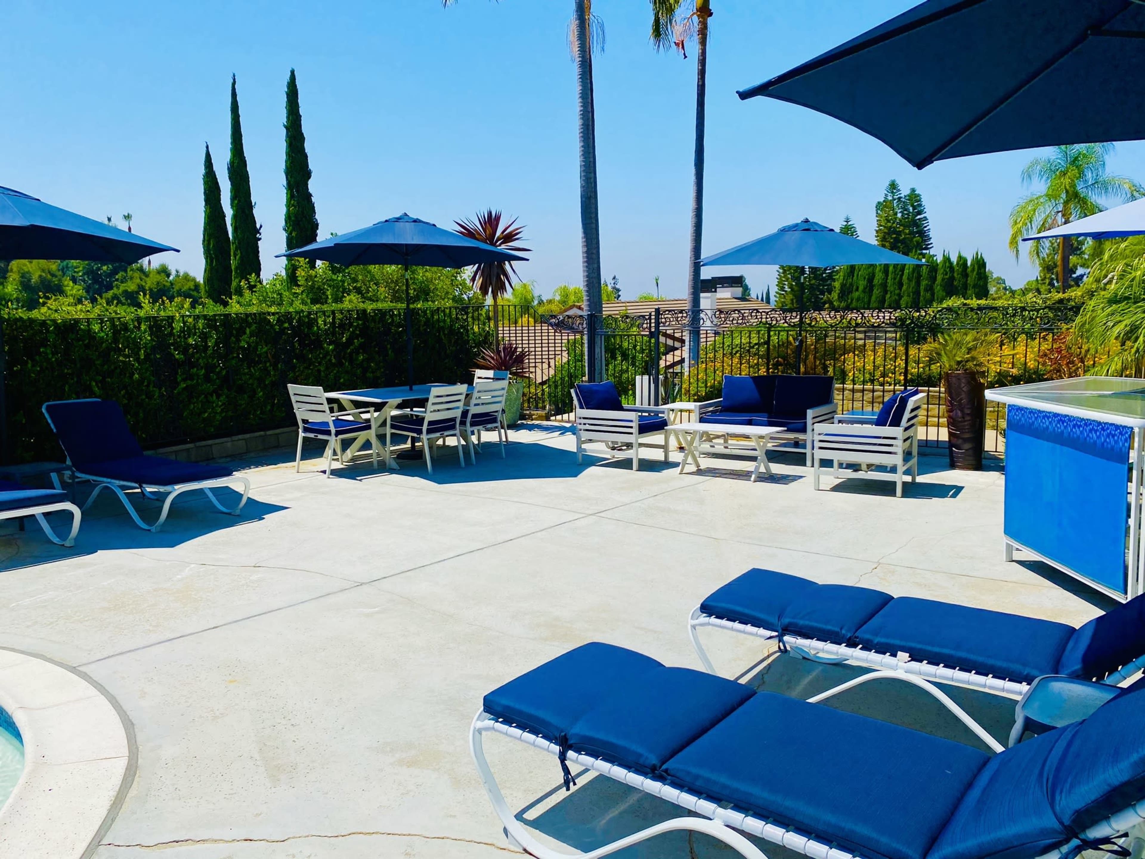 A sunny outdoor pool area with blue lounge chairs, umbrellas, and a seating arrangement surrounded by greenery.