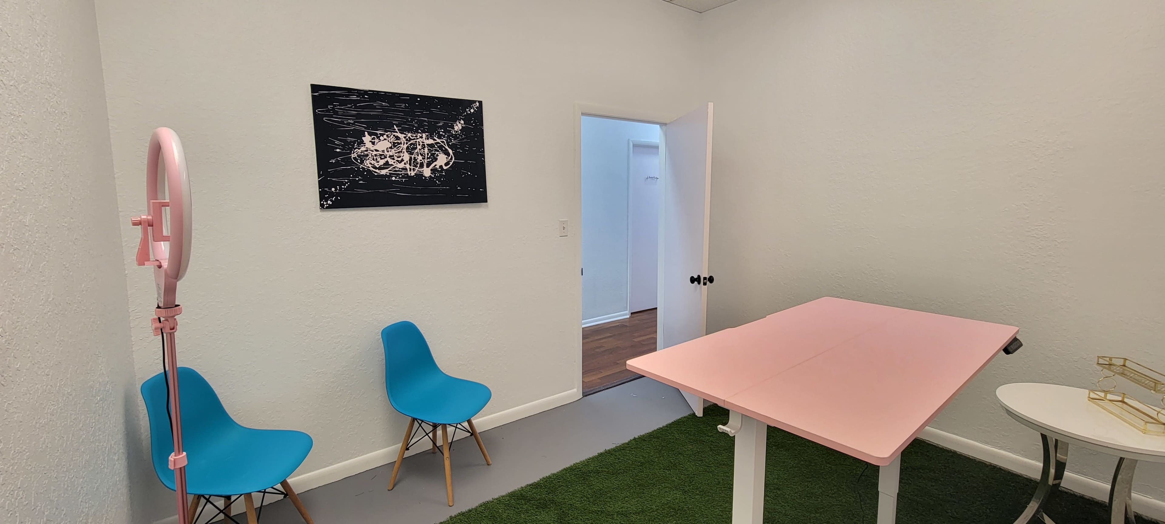 The image shows a small room with a pink table, two blue chairs, and a door leading to another area, along with a piece of art on the wall and a patch of green carpet.
