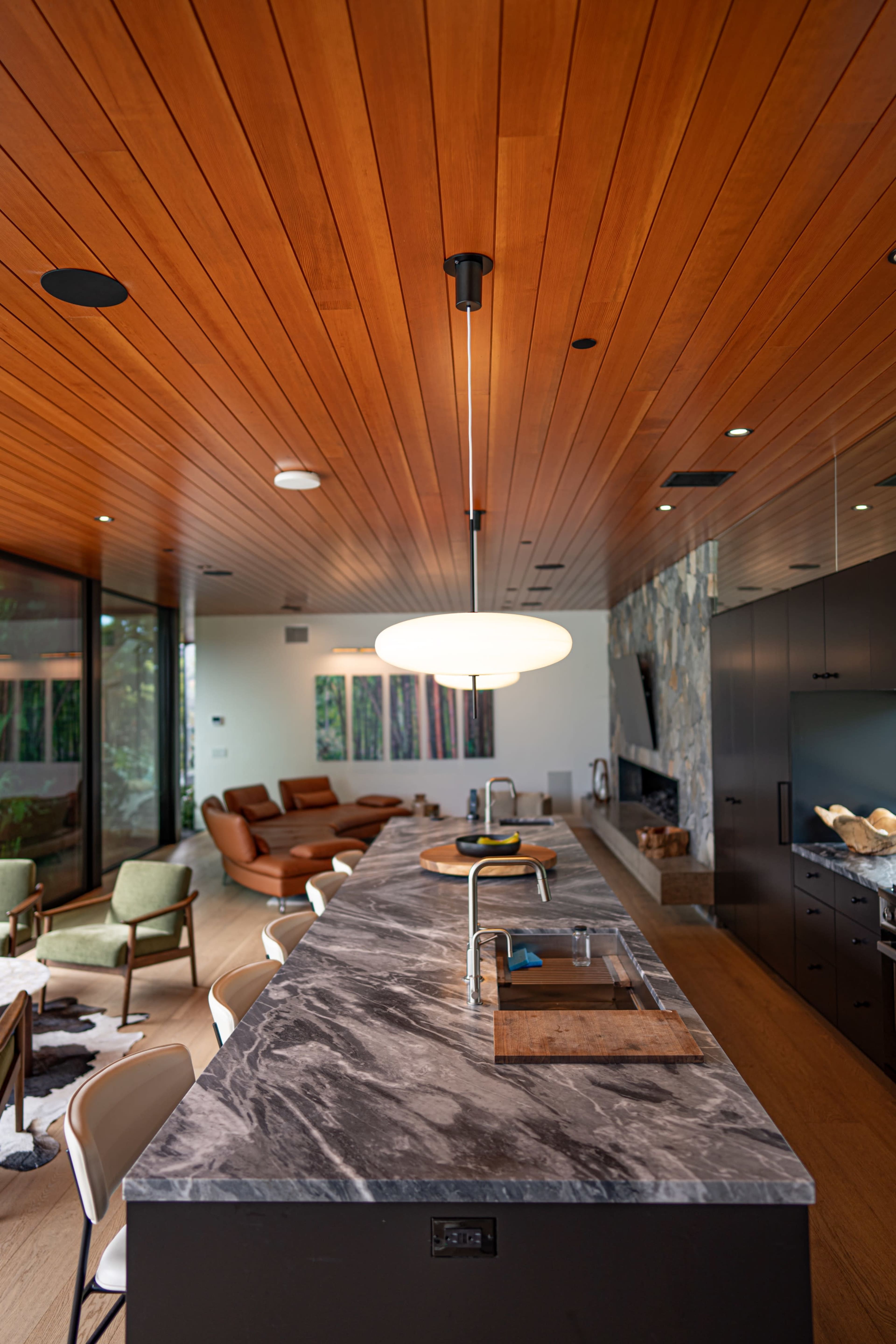 A modern kitchen with a marble countertop, wooden ceiling, and a cozy living area in the background featuring brown sofas and green chairs.